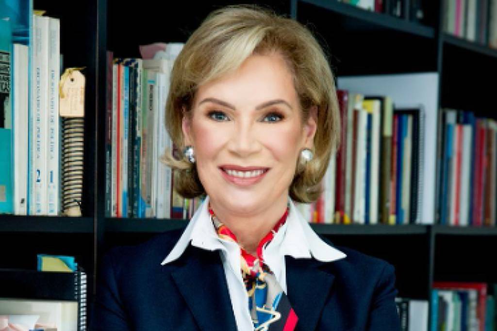 Mulher de cabelos curtos loiros veste terno azul escuro com camisa branca e lenço colorido no pescoço. Ela está com os braços cruzados, sorrindo, em frente a estantes cheias de livros.