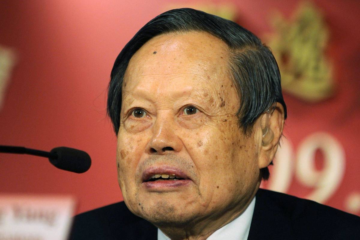 Homem asiático de idade avançada com cabelo preto penteado para o lado, veste terno escuro e camisa branca, fala em microfone diante de fundo vermelho desfocado com números e símbolos dourados.