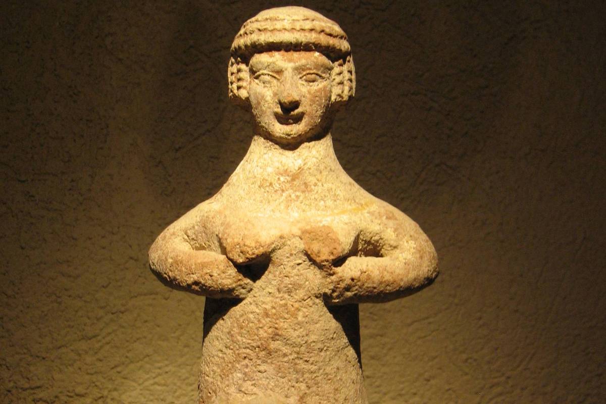 Estatueta de pedra representando figura humana com cabelo estilizado em tranças, braços curvados à frente e corpo simplificado. A peça está iluminada contra fundo neutro, exibindo textura áspera e desgaste natural.