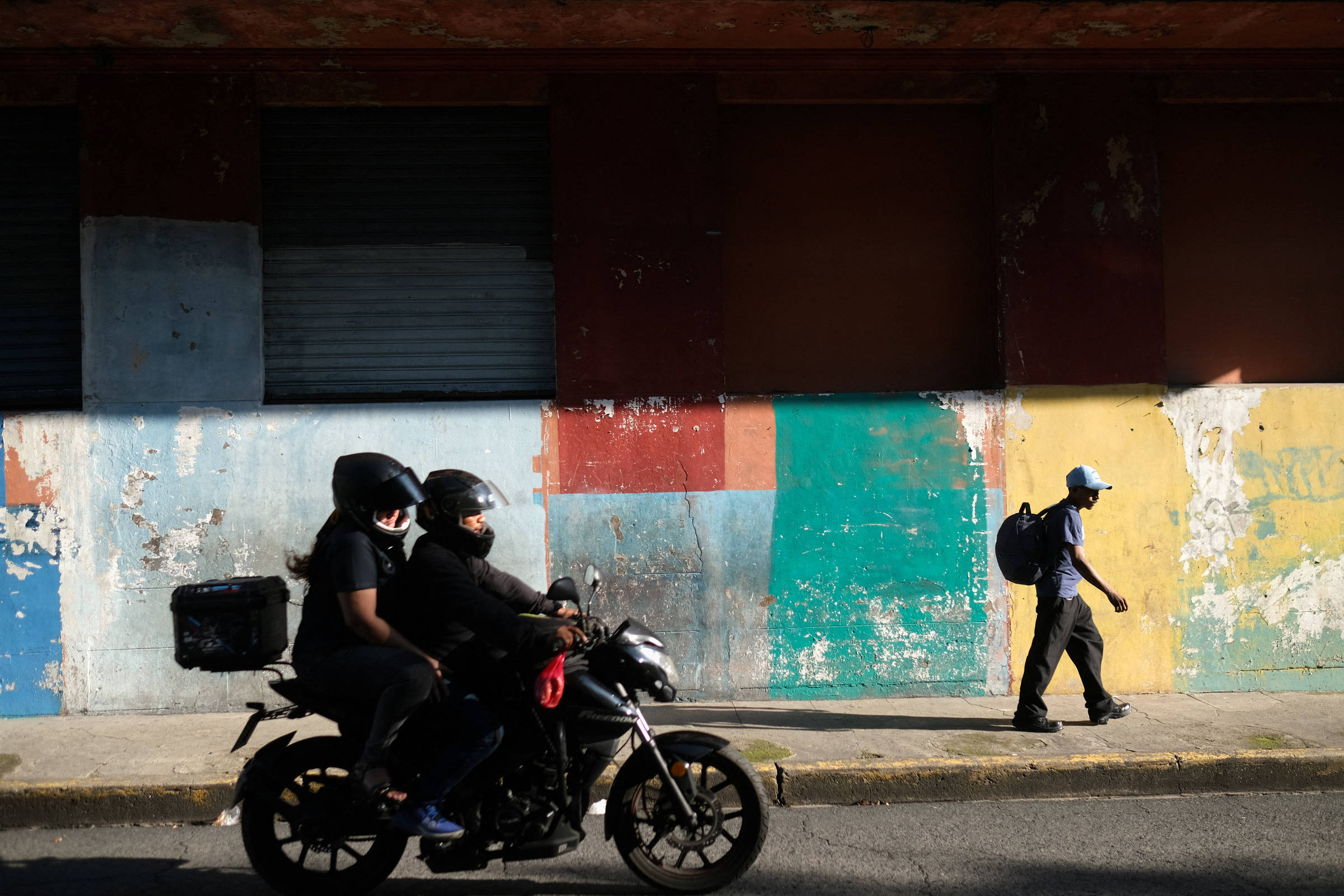 Dois motociclistas com capacetes transitam pela rua em frente a uma parede pintada com faixas nas cores azul, vermelha, verde e amarela. Um pedestre com boné e mochila caminha na calçada ao lado, sob luz do sol.