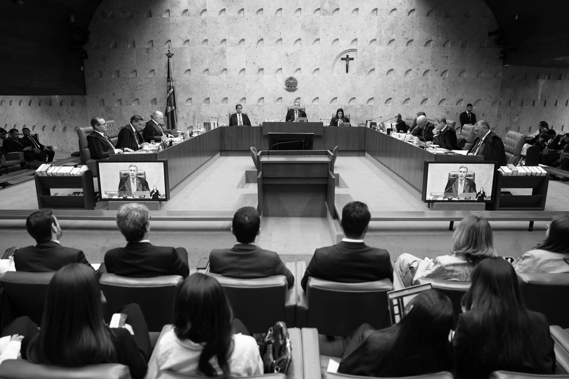 A imagem mostra uma sessão do Supremo Tribunal Federal do Brasil. No centro, há uma mesa retangular com vários ministros sentados, alguns olhando para a tela à sua frente. À esquerda, há uma bandeira do Brasil e, ao fundo, uma cruz. Na parte inferior da imagem, é possível ver um grupo de pessoas sentadas, algumas com laptops e outras com anotações, assistindo à sessão. As paredes são de um tom claro e texturizadas.
