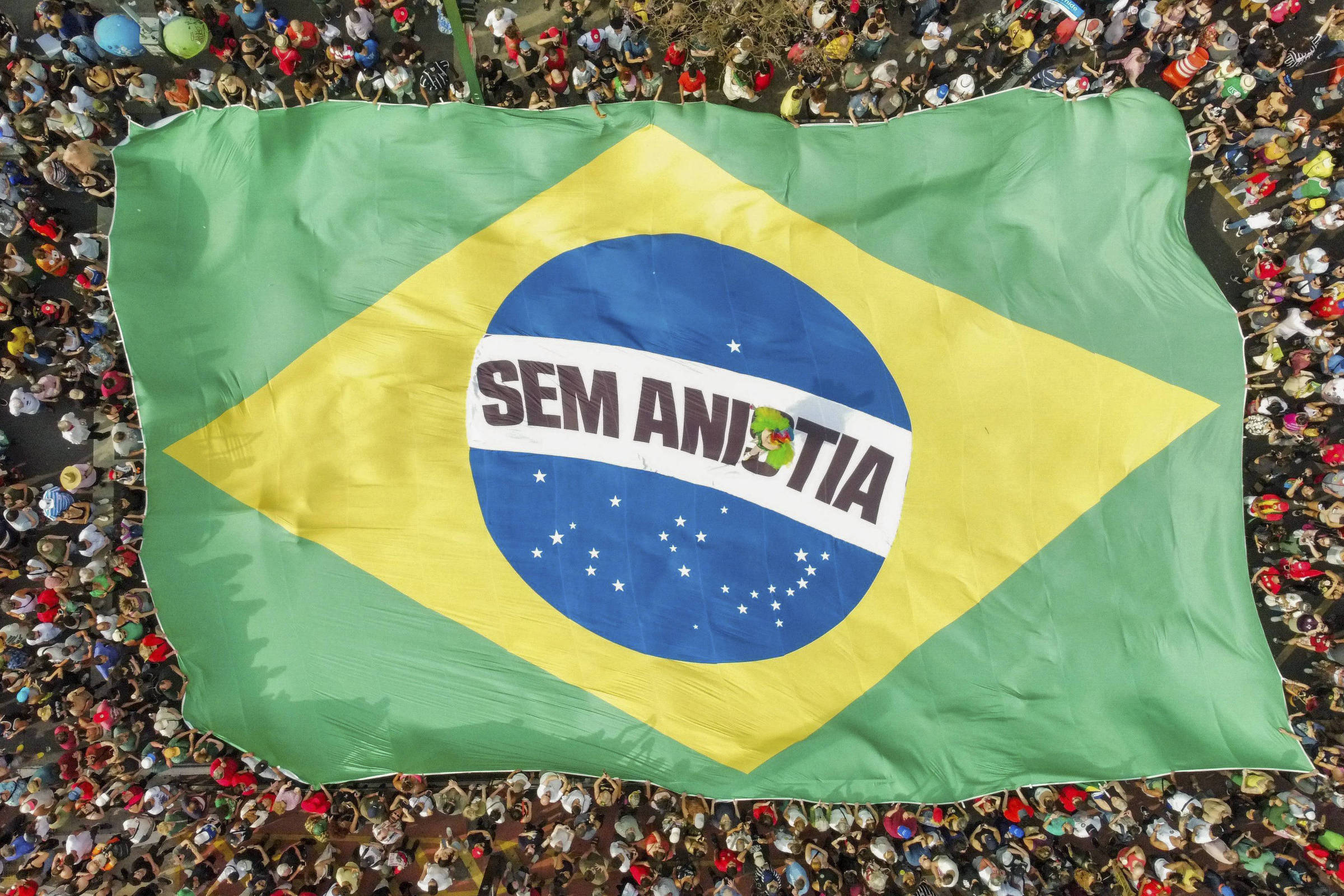 Multidão vista de cima rodeia uma grande bandeira do Brasil estendida no chão. No centro da bandeira, onde normalmente está a faixa com o lema nacional, há uma faixa branca com a palavra