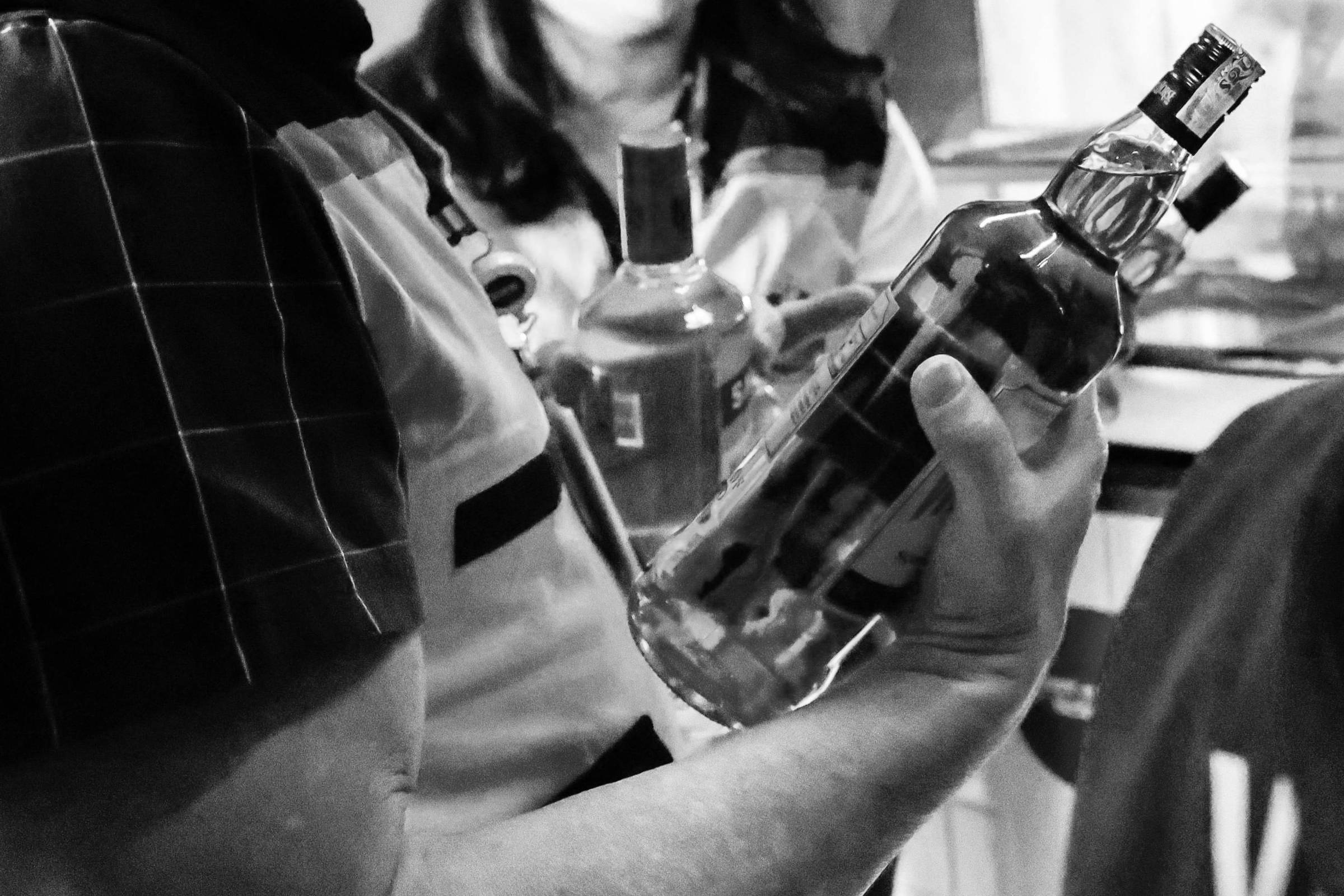 Agente segura garrafa de bebida alcoólica com rótulo laranja enquanto outra pessoa ao fundo segura garrafa com tampa vermelha. Ambos usam coletes amarelos, indicando atividade de fiscalização.