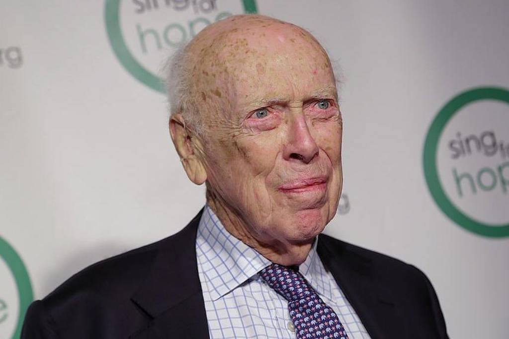 Homem idoso com terno preto, camisa xadrez branca e gravata roxa posando em frente a fundo branco com logotipo verde e texto