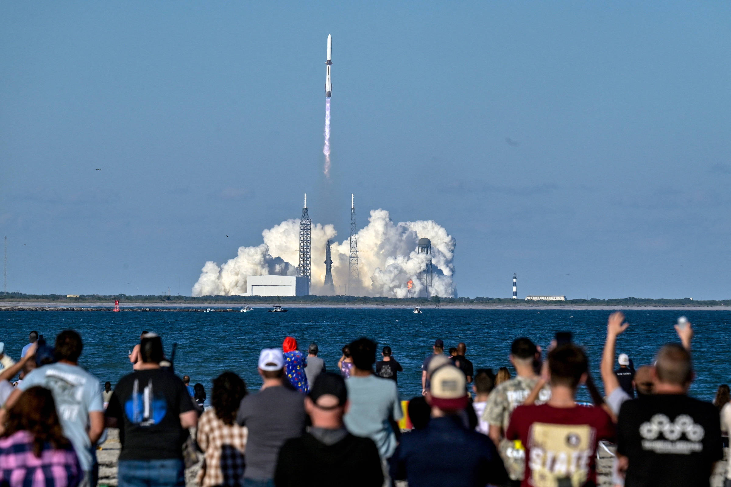 Foguete decolando com chamas e fumaça densa na base, visto ao longe sobre o mar. Pessoas na costa assistem e registram o lançamento em dia claro.