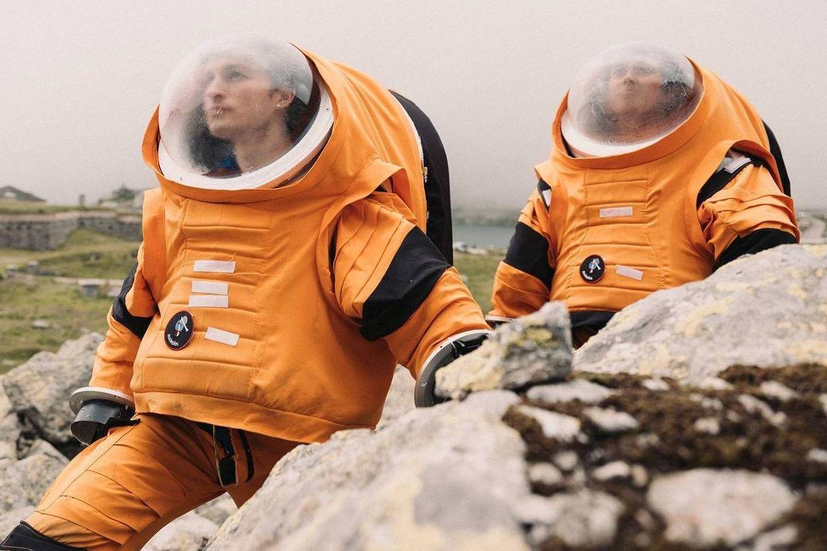 Duas pessoas vestem trajes espaciais laranja com capacetes transparentes, posicionadas em terreno rochoso e gramado. O céu está nublado e o ambiente parece ao ar livre, possivelmente em simulação ou treinamento.