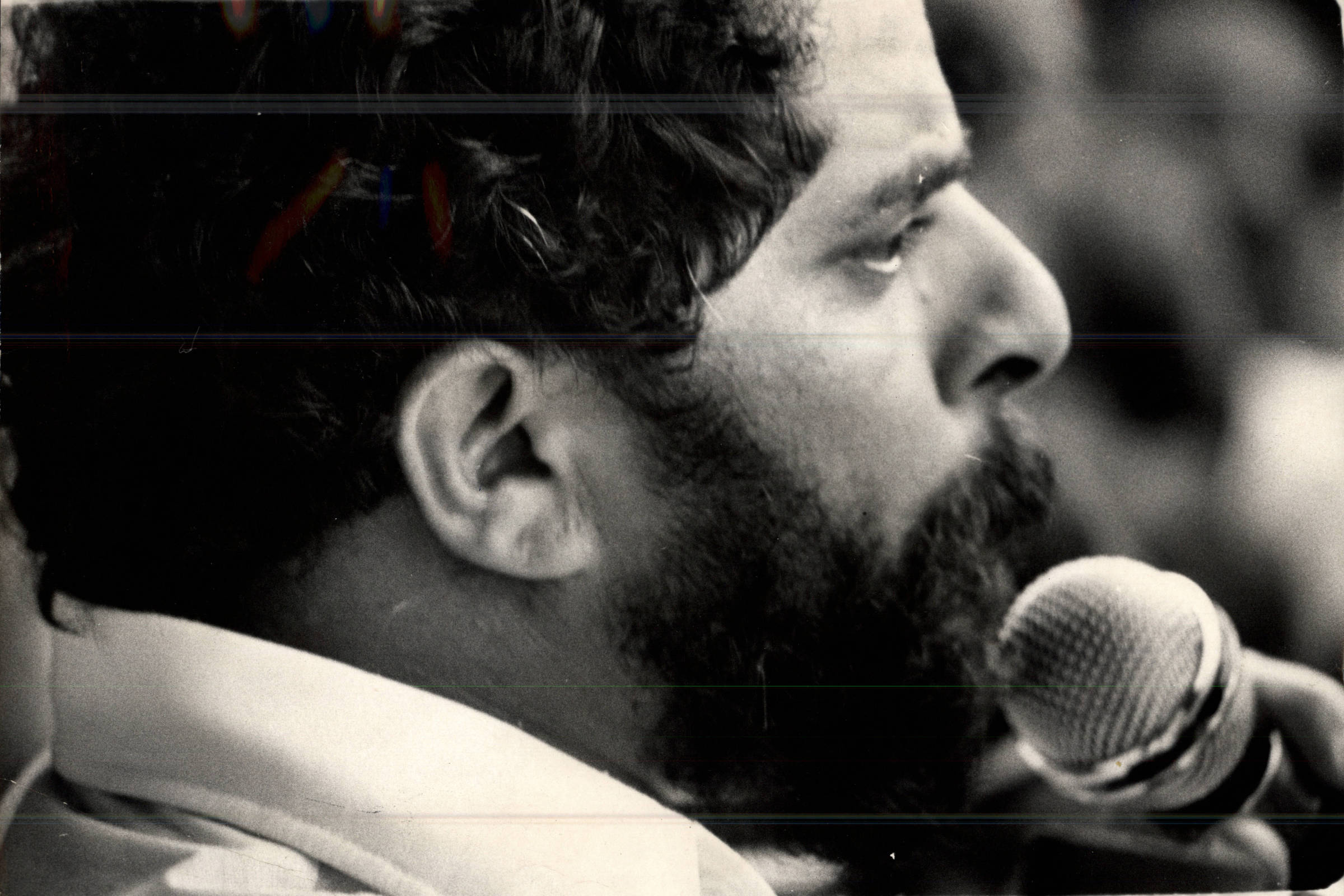Retrato em preto e branco mostra homem de perfil com barba e cabelo encaracolado, falando ao microfone próximo à boca. Fundo desfocado sugere ambiente interno.