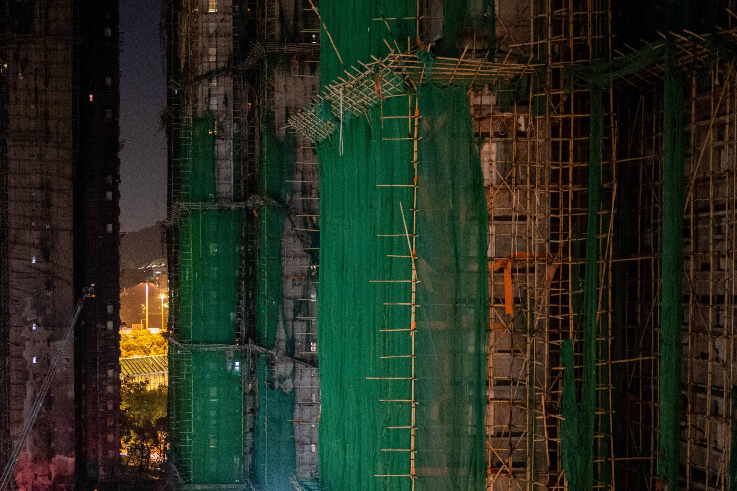 Prédios altos em construção com estruturas de andaimes e telas verdes cobrindo as fachadas. Iluminação artificial destaca detalhes das obras durante a noite.