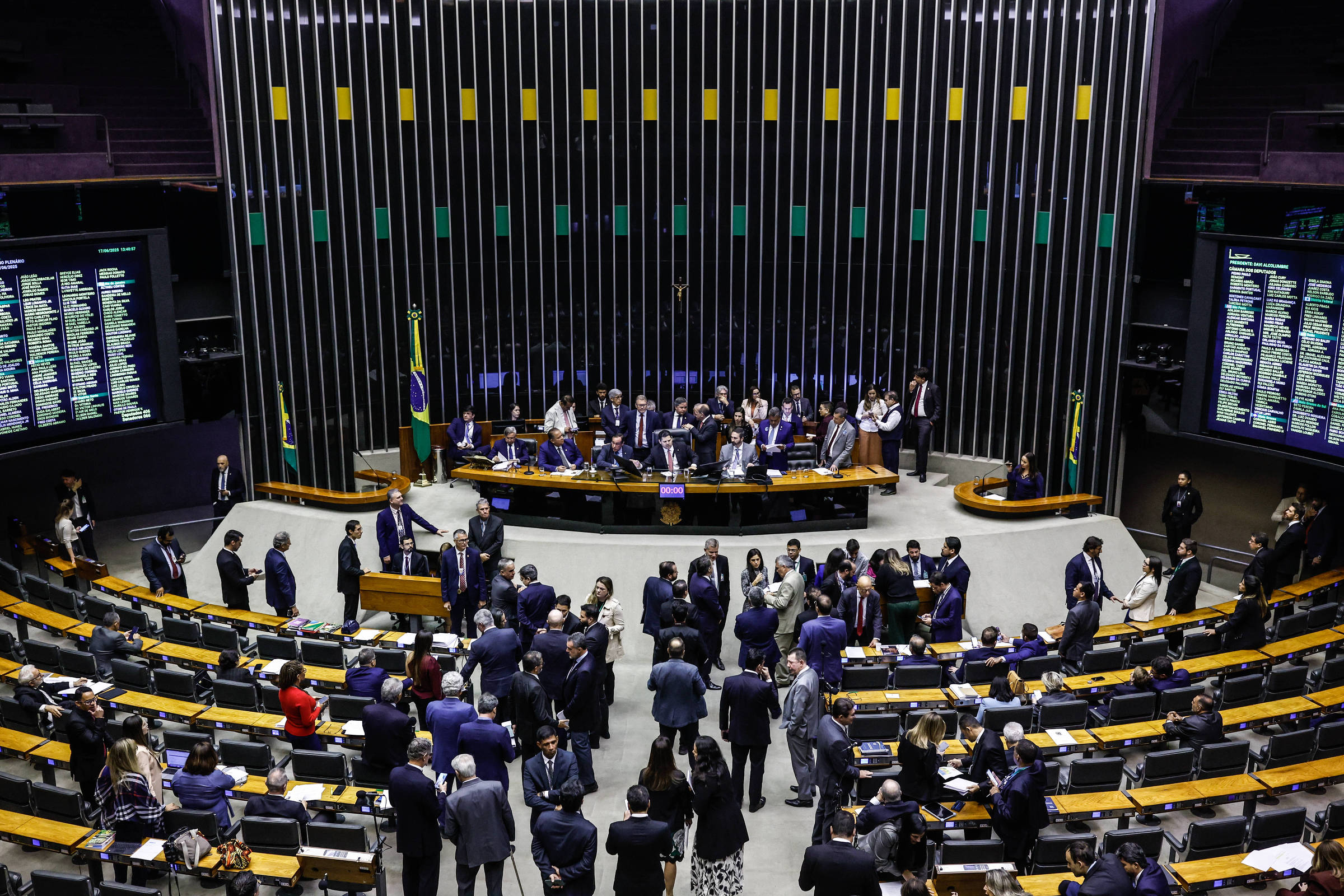 A imagem mostra o plenário da Câmara dos Deputados do Brasil, com várias pessoas em trajes formais interagindo. No fundo, há uma mesa com deputados e bandeiras do Brasil. As paredes são decoradas com painéis e telas que exibem informações. O ambiente é bem iluminado e moderno, com assentos dispostos em fileiras