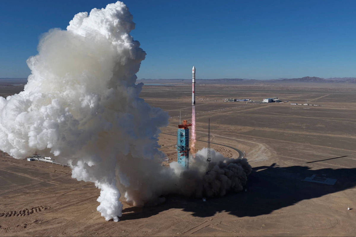Foguete decolando em plataforma no deserto, com grande nuvem de fumaça branca e poeira ao redor. Céu azul claro e terreno árido ao fundo.