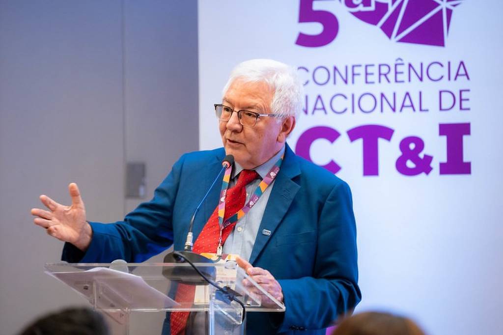 Homem idoso de cabelos brancos e óculos fala em púlpito transparente durante a 5ª Conferência Nacional de CT&I. Ele veste terno azul, camisa clara e gravata vermelha com padrão colorido, e gesticula com a mão esquerda.