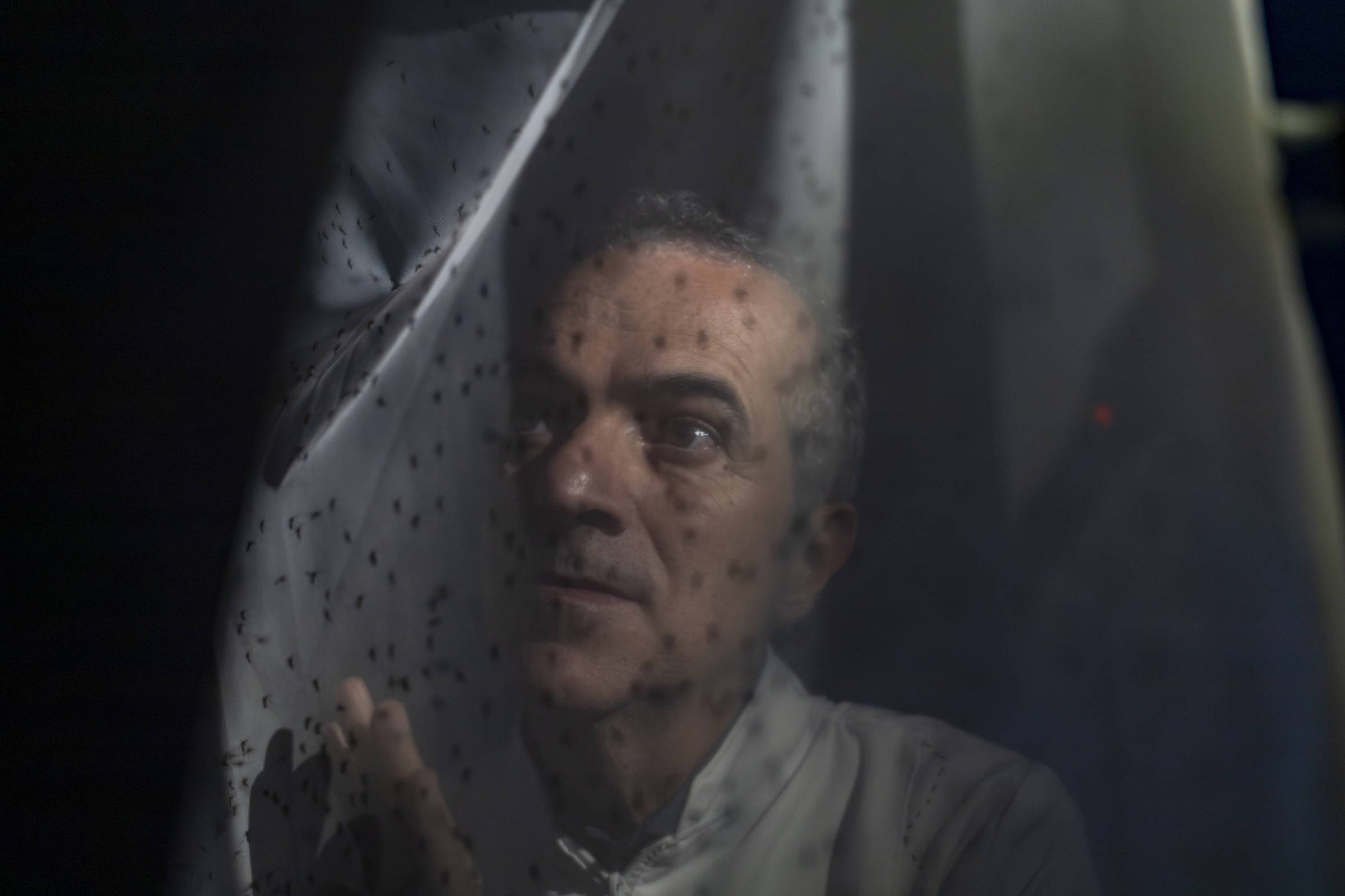 Homem de meia-idade com expressão séria observa através de uma cortina translúcida com pequenos pontos escuros. A iluminação é baixa, destacando seu rosto parcialmente iluminado e o tecido da cortina.