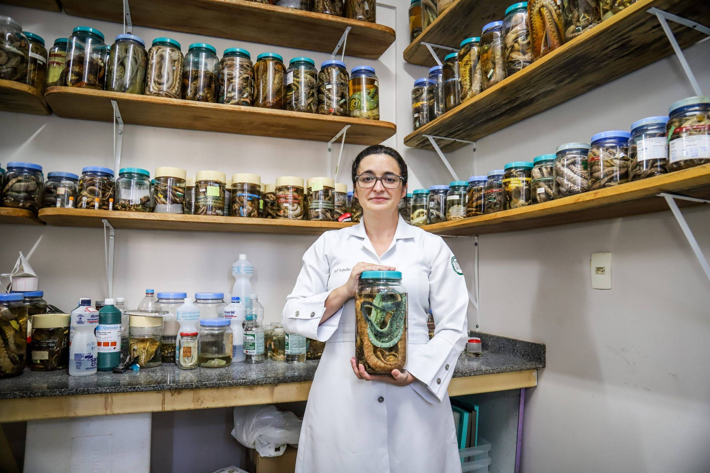 Mulher com jaleco branco segura um grande frasco de vidro contendo espécimes preservados em líquido. Ela está em um laboratório com prateleiras ao fundo cheias de frascos semelhantes organizados.