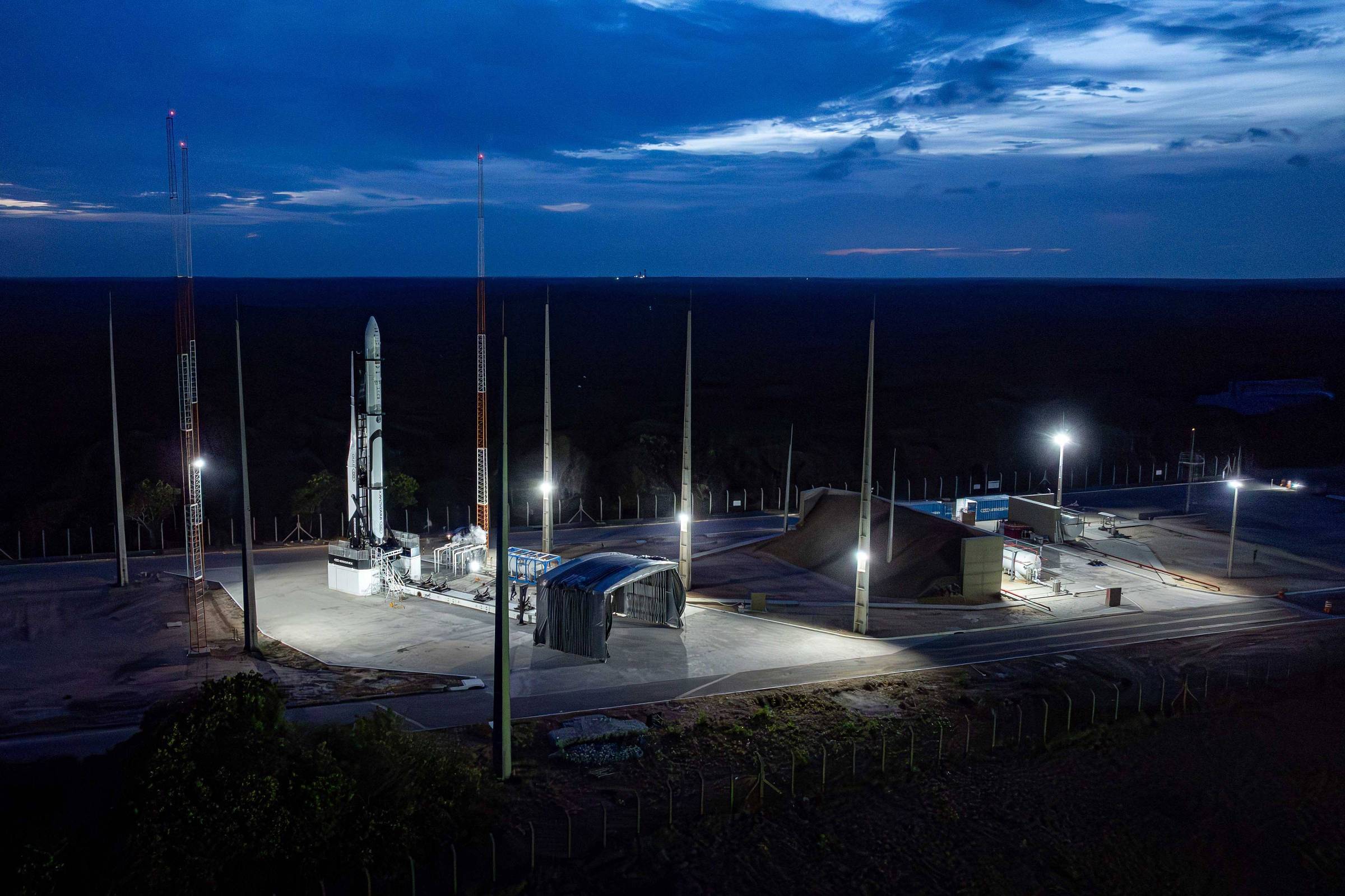 Foguete branco posicionado em plataforma de lançamento iluminada por holofotes, cercada por torres altas e vegetação ao fundo sob céu azul escuro com nuvens.