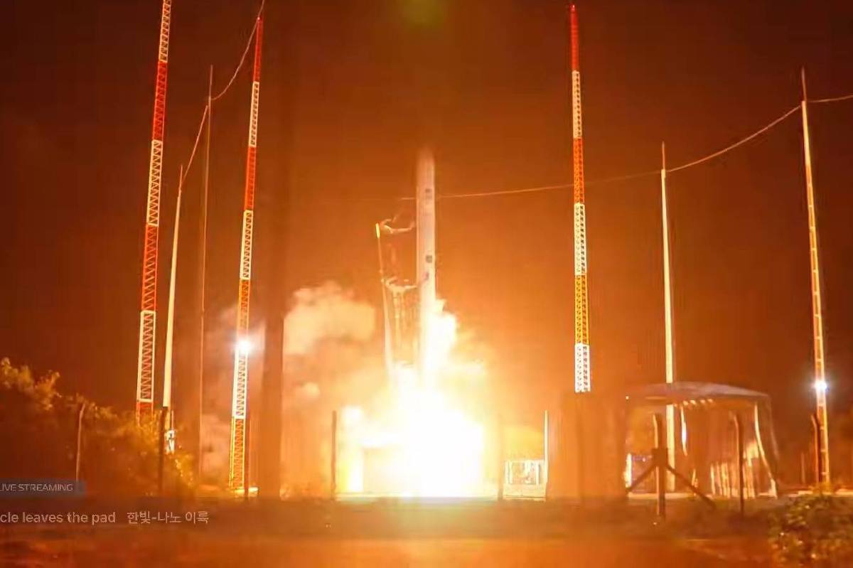 Foguete decolando à noite no Centro Espacial de Alcântara, com chamas intensas e fumaça na base. Torres de suporte iluminadas cercam a plataforma de lançamento.