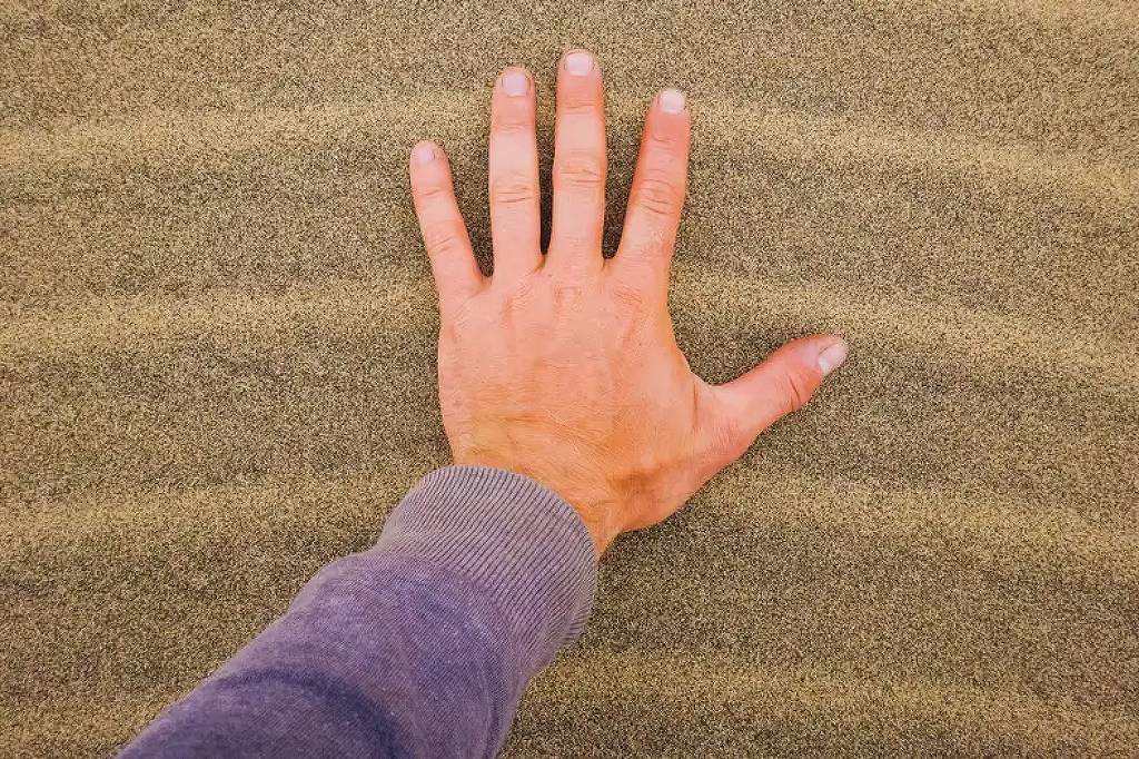 Mão direita de pessoa com manga roxa apoiada sobre areia fina com ondulações paralelas.