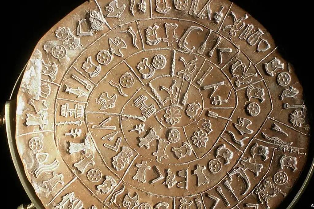 Disco de argila circular com inscrições em espiral contendo símbolos pictográficos variados, incluindo figuras humanas, animais e objetos, exibido em suporte metálico.