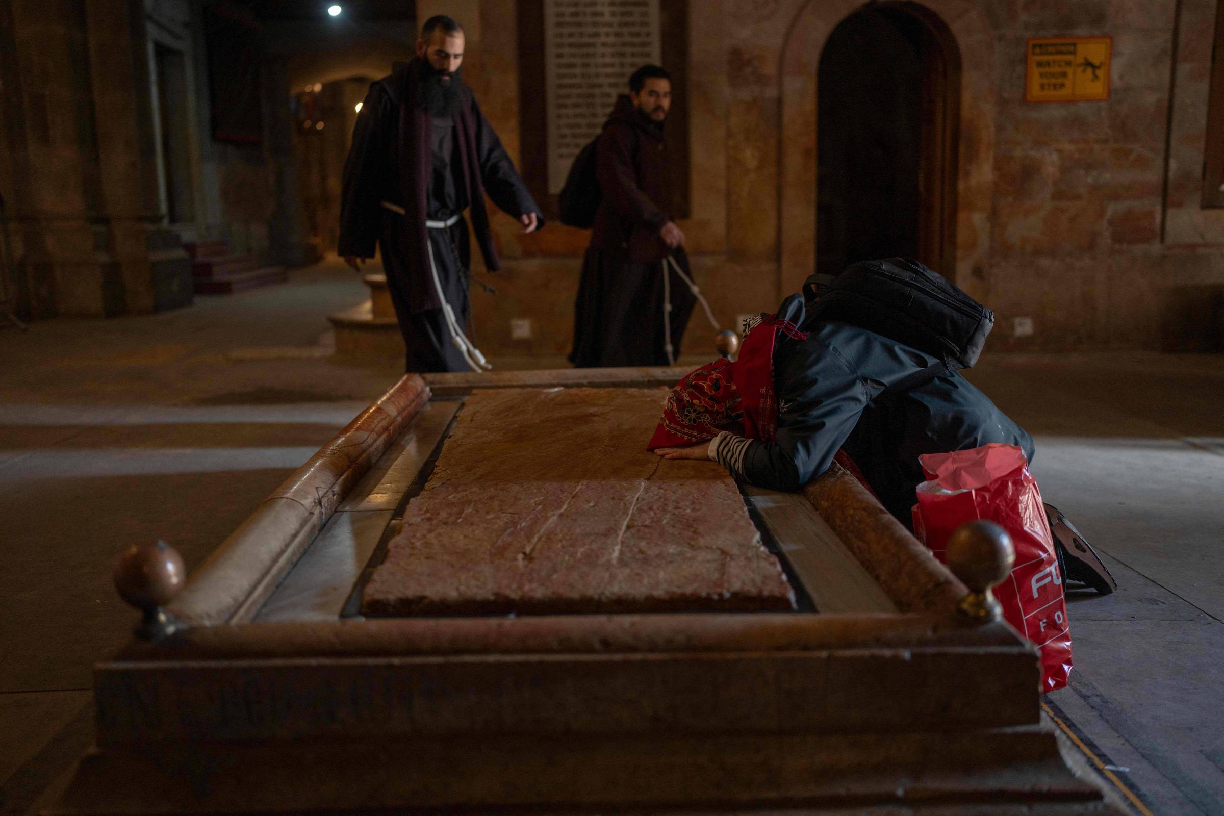 Pessoa ajoelhada com cabeça sobre túmulo de pedra em interior de construção antiga. Dois monges vestidos com hábitos escuros caminham ao fundo próximo a paredes de pedra e arcos. Iluminação baixa destaca o túmulo e a figura ajoelhada.