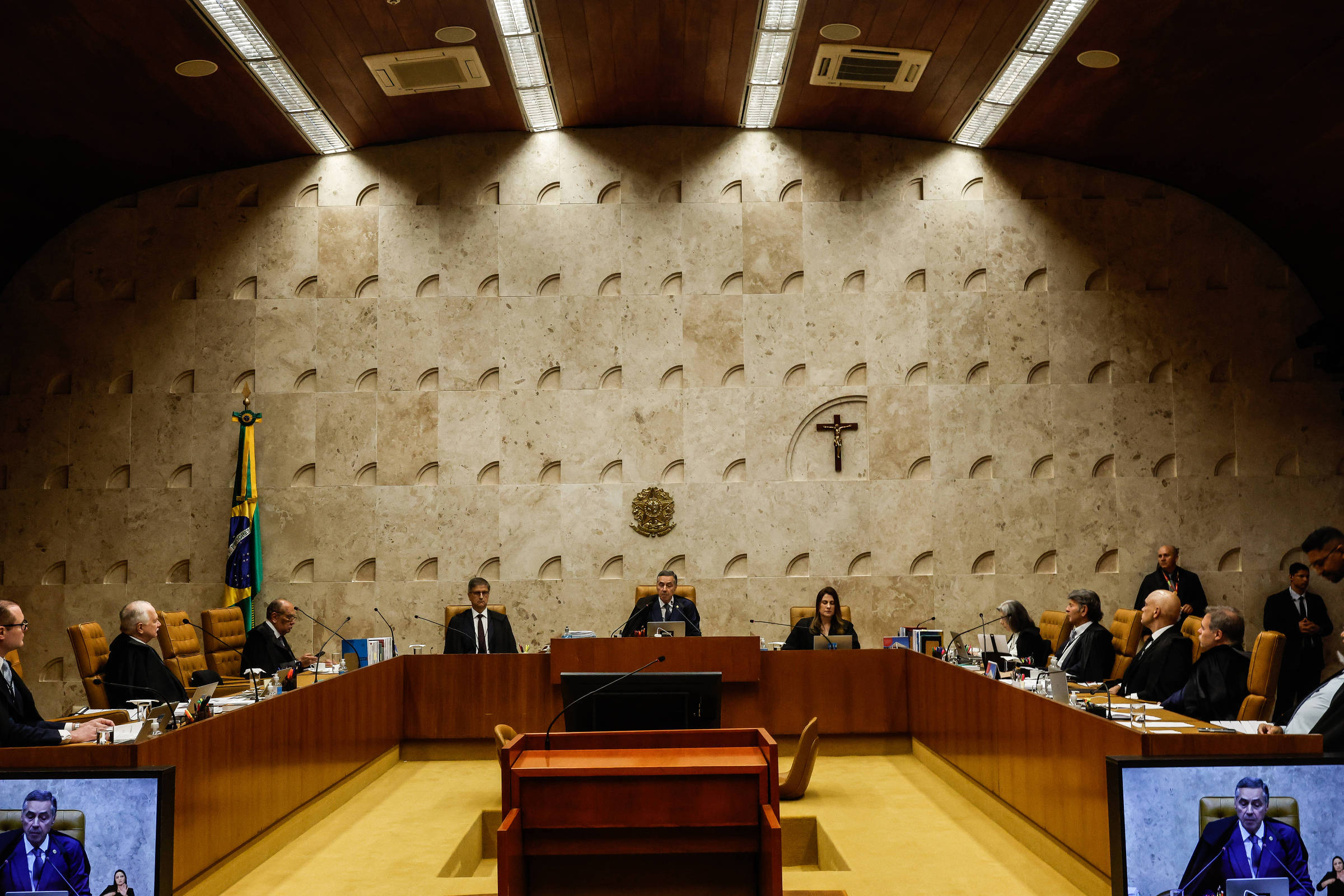 A imagem mostra uma sessão do Supremo Tribunal Federal do Brasil. O ambiente é formal, com uma mesa em formato retangular onde estão sentados vários ministros. Ao fundo, há uma bandeira do Brasil e um símbolo de cruz. A iluminação é suave e as paredes têm um design texturizado. Na frente, há telas que exibem um orador