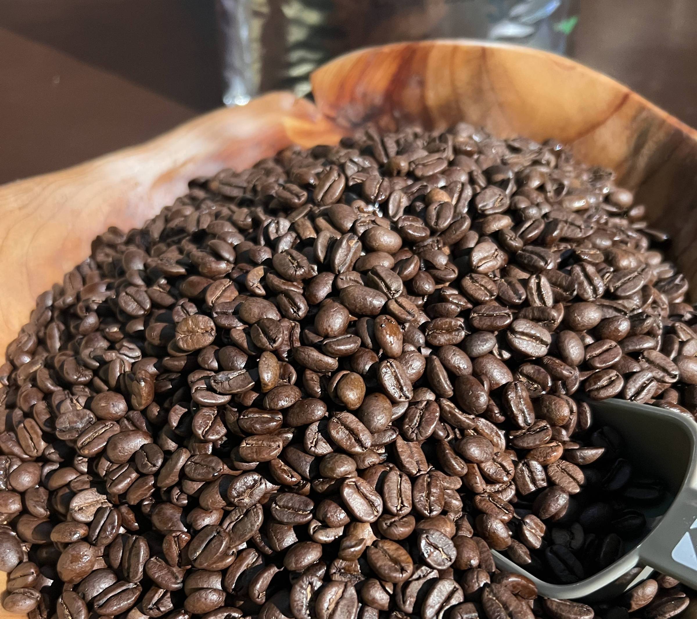 Tigela de madeira cheia de grãos de café torrados escuros com uma colher cinza parcialmente enterrada entre os grãos.