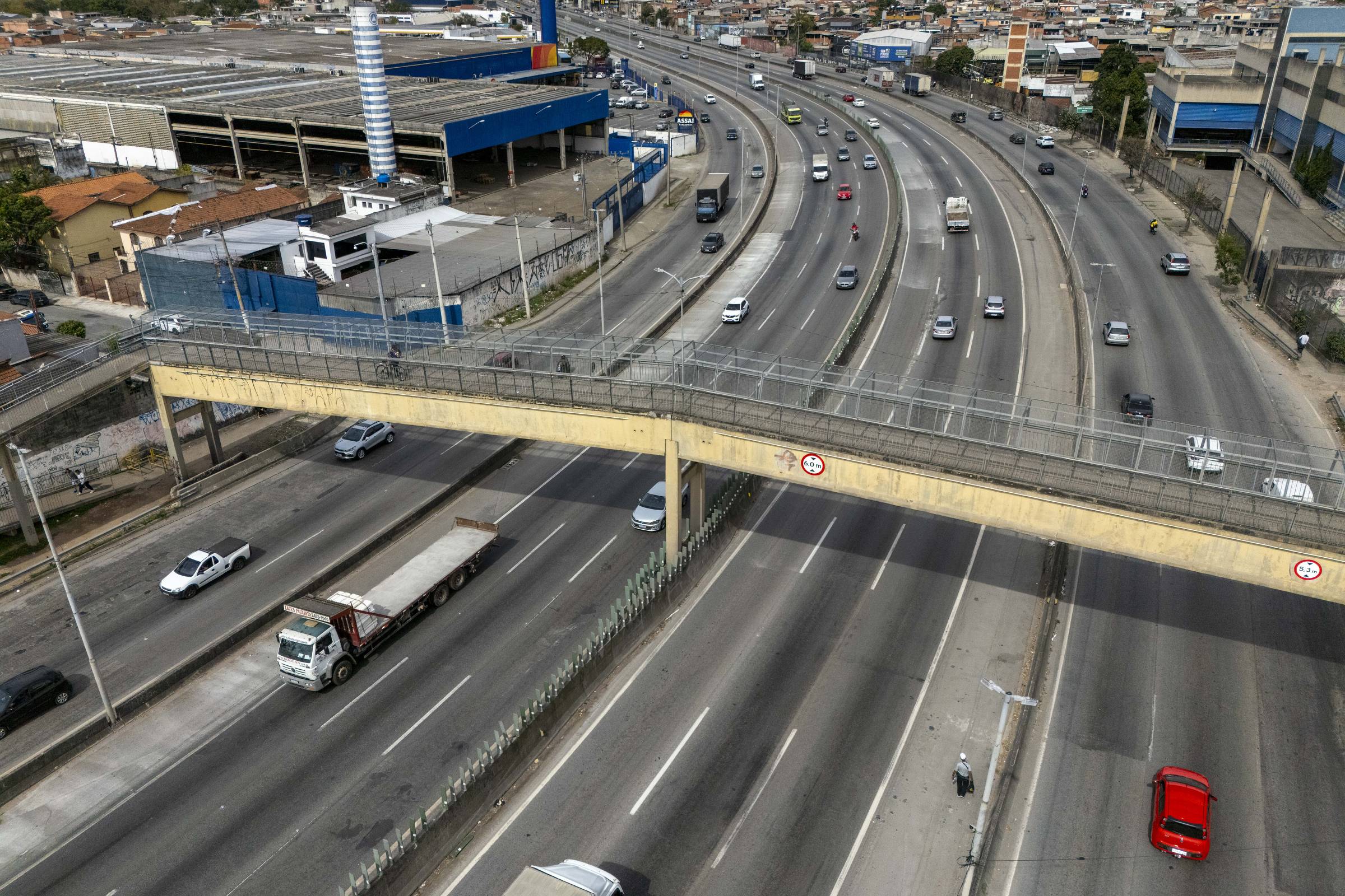 A imagem mostra uma rodovia com várias faixas de tráfego. Há veículos em movimento, incluindo caminhões e carros. No primeiro plano, uma passarela para pedestres se destaca, conectando os dois lados da rodovia. Ao fundo, é possível ver um grande edifício e áreas urbanas. O céu está parcialmente nublado.