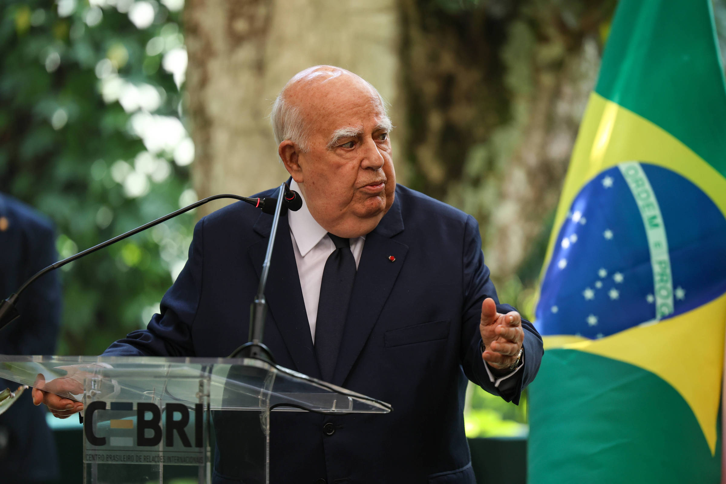 Um homem idoso, com cabelo grisalho e usando um terno escuro, está falando em um microfone. Ele gesticula com a mão direita enquanto se dirige a uma plateia. Ao fundo, há uma bandeira do Brasil e uma estrutura de vidro com a inscrição