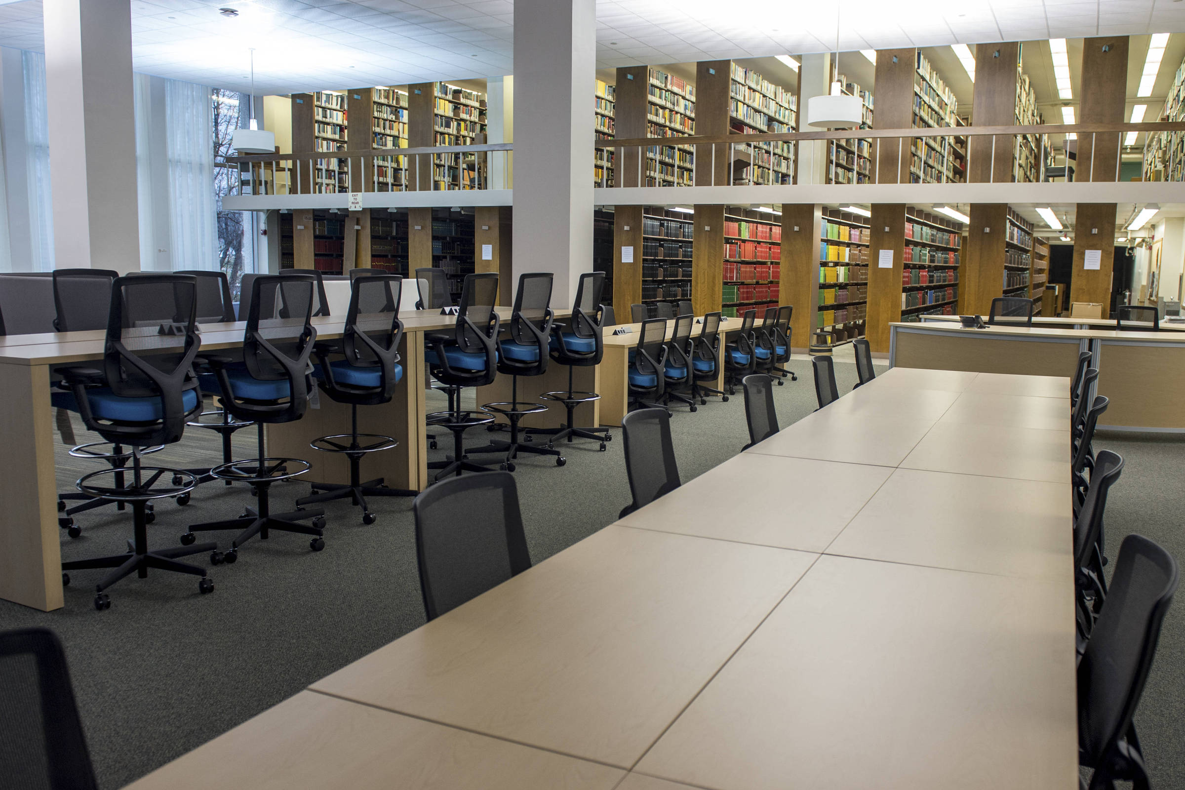 Área de estudo em biblioteca com longas mesas claras e cadeiras pretas alinhadas. Ao fundo, estantes altas cheias de livros organizados em dois andares, com cadeiras altas em balcões para leitura individual.