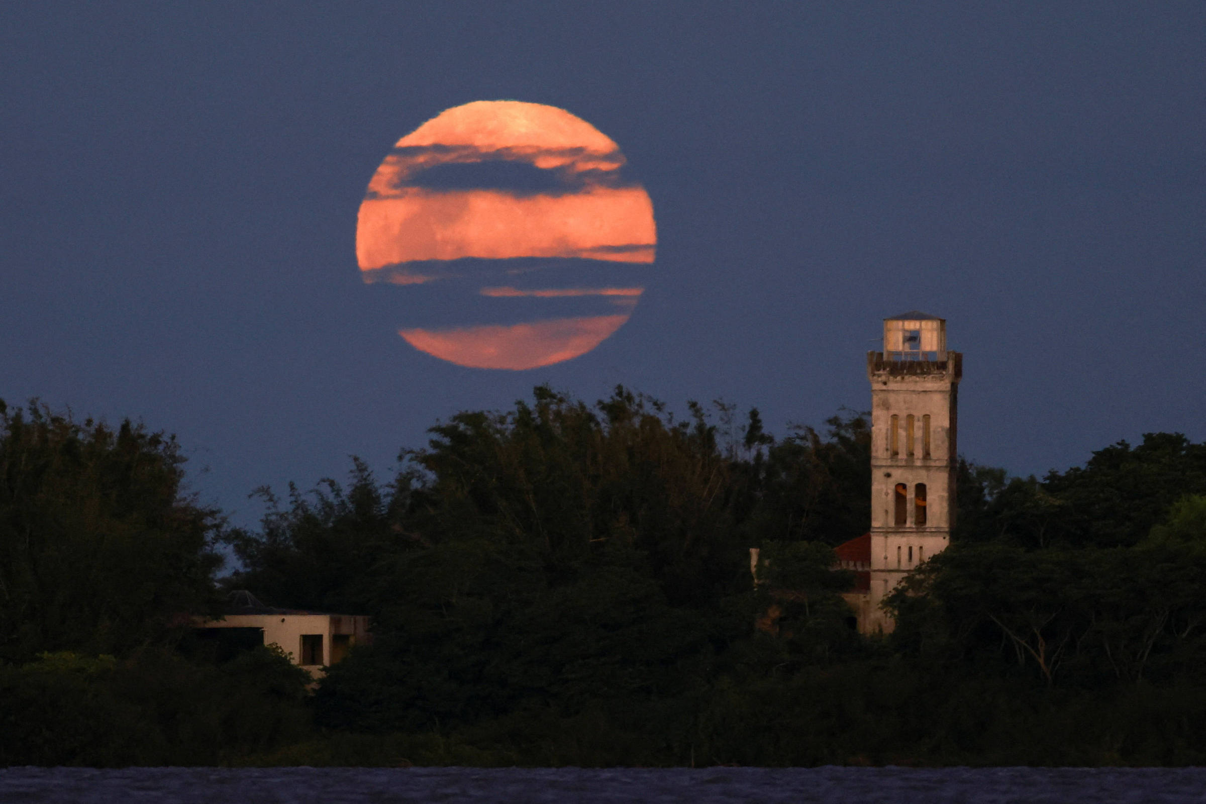 Lua cheia de tom alaranjado aparece parcialmente encoberta por nuvens escuras no céu noturno. À direita, torre antiga de pedra se destaca entre árvores escuras na linha do horizonte.