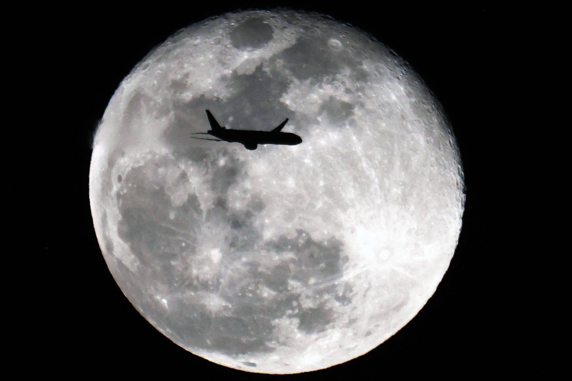 Silhueta de um avião cruzando o céu noturno com a lua cheia ao fundo, destacando detalhes da superfície lunar.