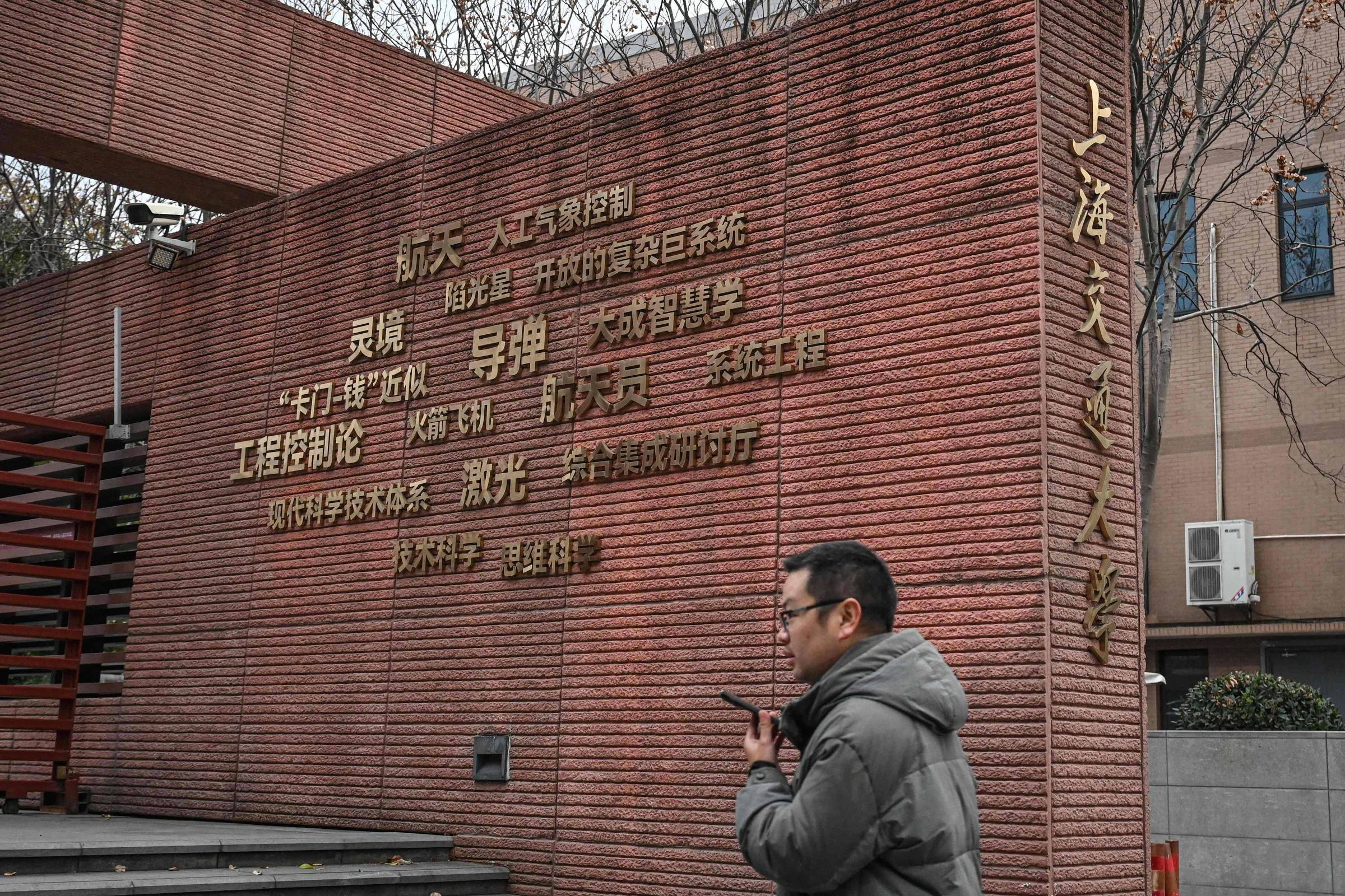 Homem de casaco cinza caminha segurando celular em frente a muro de tijolos vermelhos com texto em chinês dourado. Ambiente urbano com árvores sem folhas ao fundo.