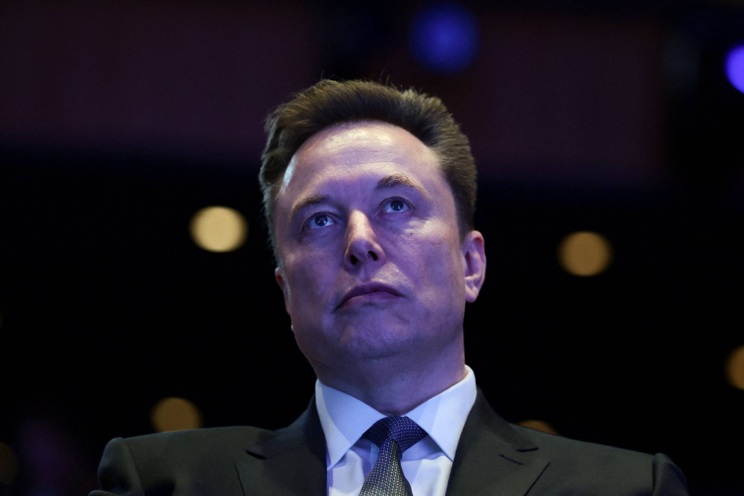 Homem de meia-idade com cabelo curto e escuro veste terno preto, camisa branca e gravata cinza, olhando levemente para cima em ambiente escuro com luzes desfocadas ao fundo.