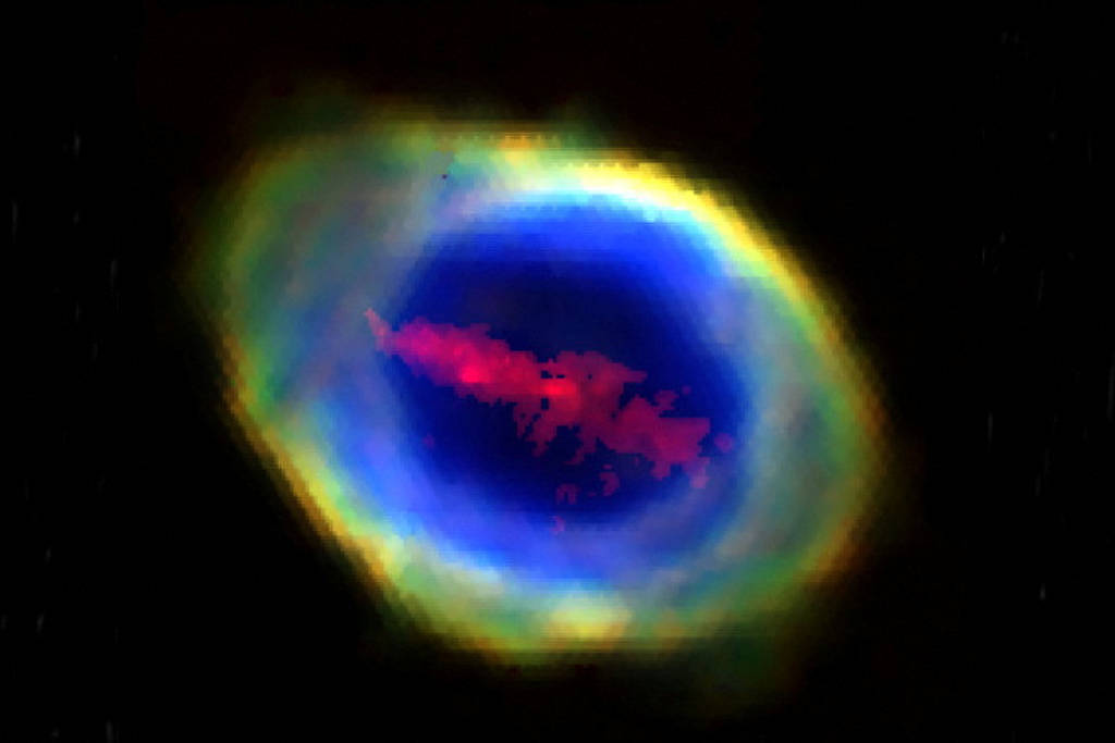 Nebulosa planetária com núcleo azul intenso cercado por anel difuso em tons de verde, amarelo e vermelho. Fundo preto com pontos de estrelas distantes.