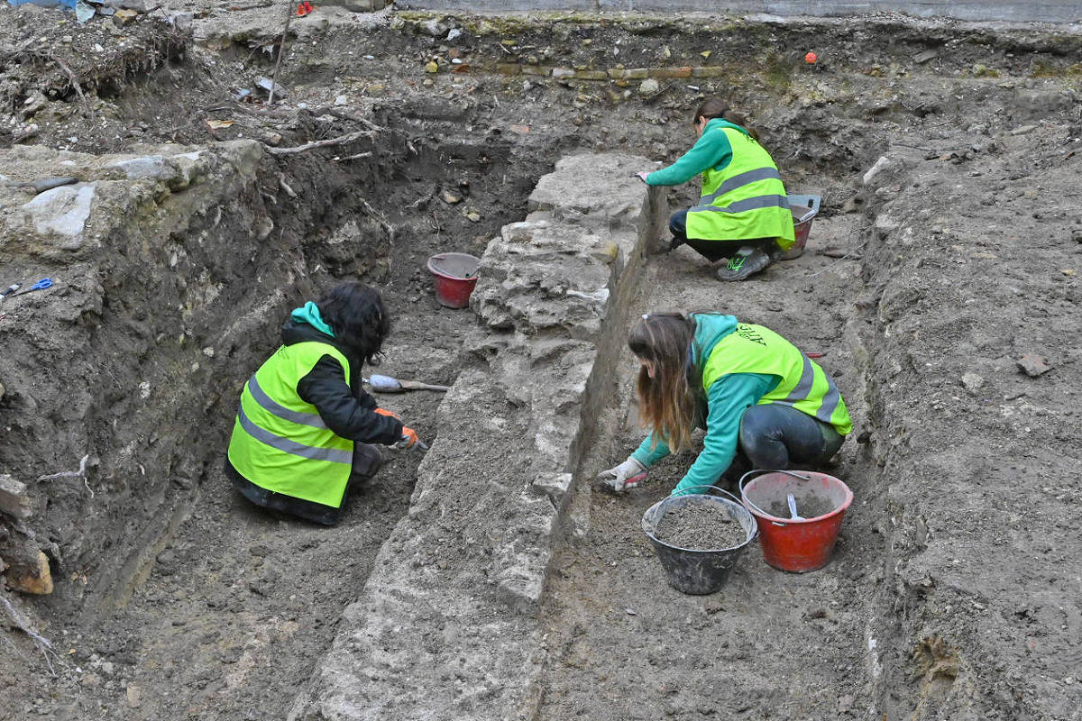 Três pessoas com coletes amarelos realizam escavação arqueológica em área de terra. Duas estão agachadas limpando uma estrutura de pedra exposta, enquanto a terceira está sentada no solo próximo. Baldes vermelhos com terra e ferramentas estão ao redor.