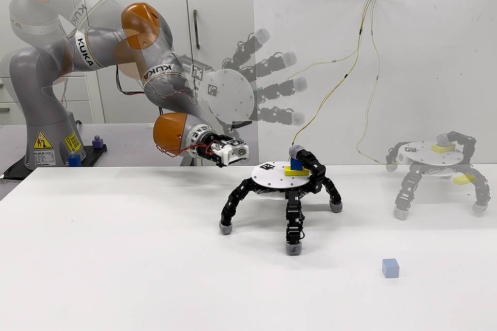 Braço robótico cinza e laranja manipula um pequeno cubo azul sobre mesa branca. Robô de três pernas está na mesa, com imagem fantasma indicando movimento para a direita. Fundo branco com paredes e armários.