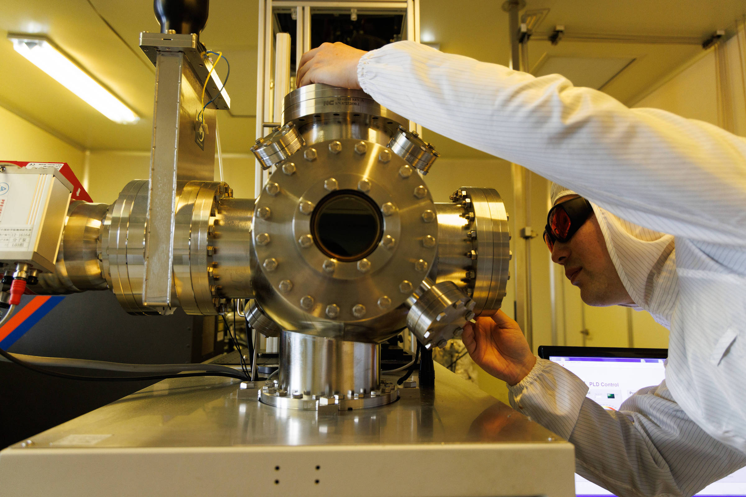 Pessoa vestindo roupa protetora branca e óculos de segurança ajusta componente de equipamento metálico complexo em laboratório. Máquina possui múltiplas conexões cilíndricas e parafusos visíveis.