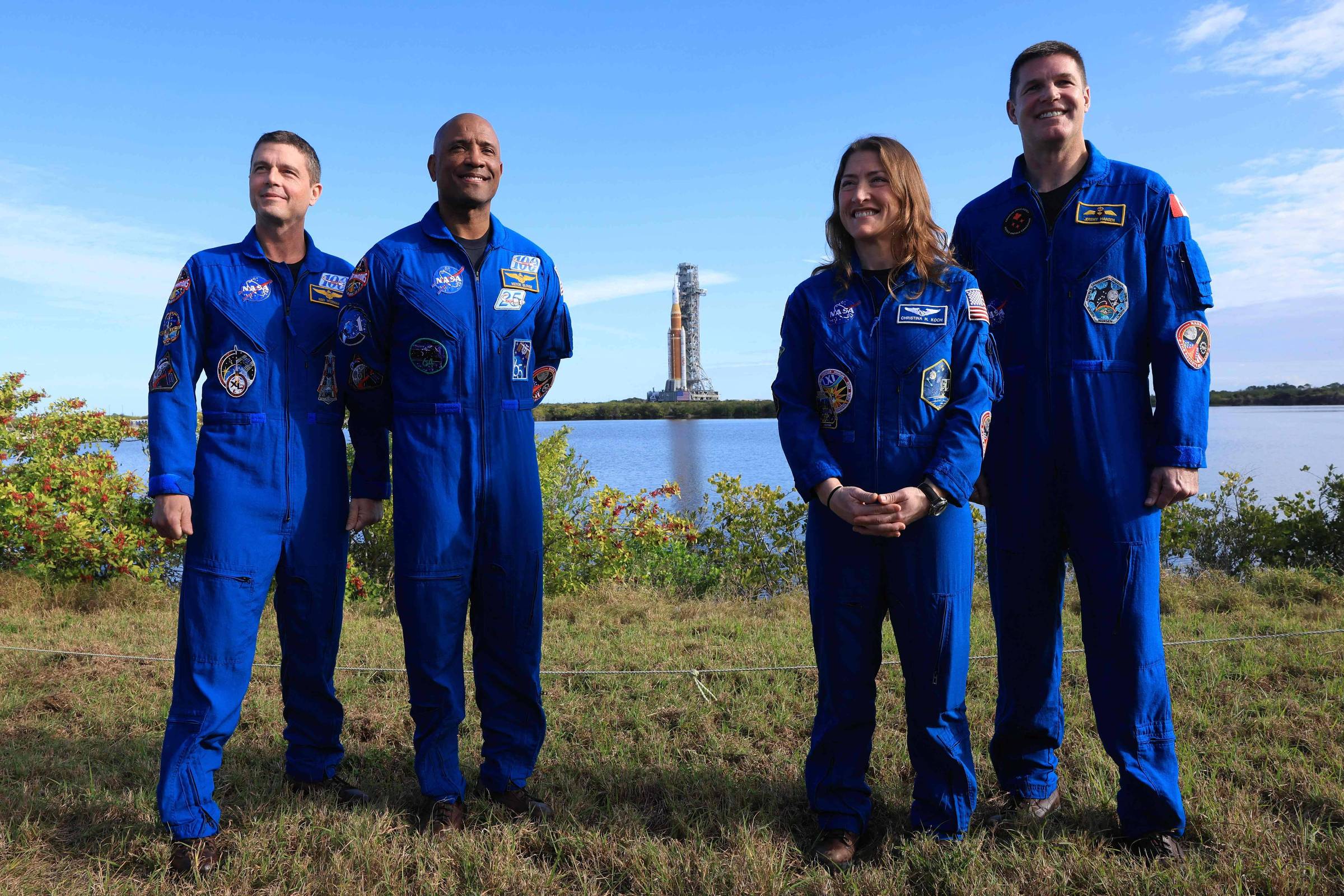 Quatro astronautas em trajes azuis posam em pé sobre grama com vegetação e corpo d