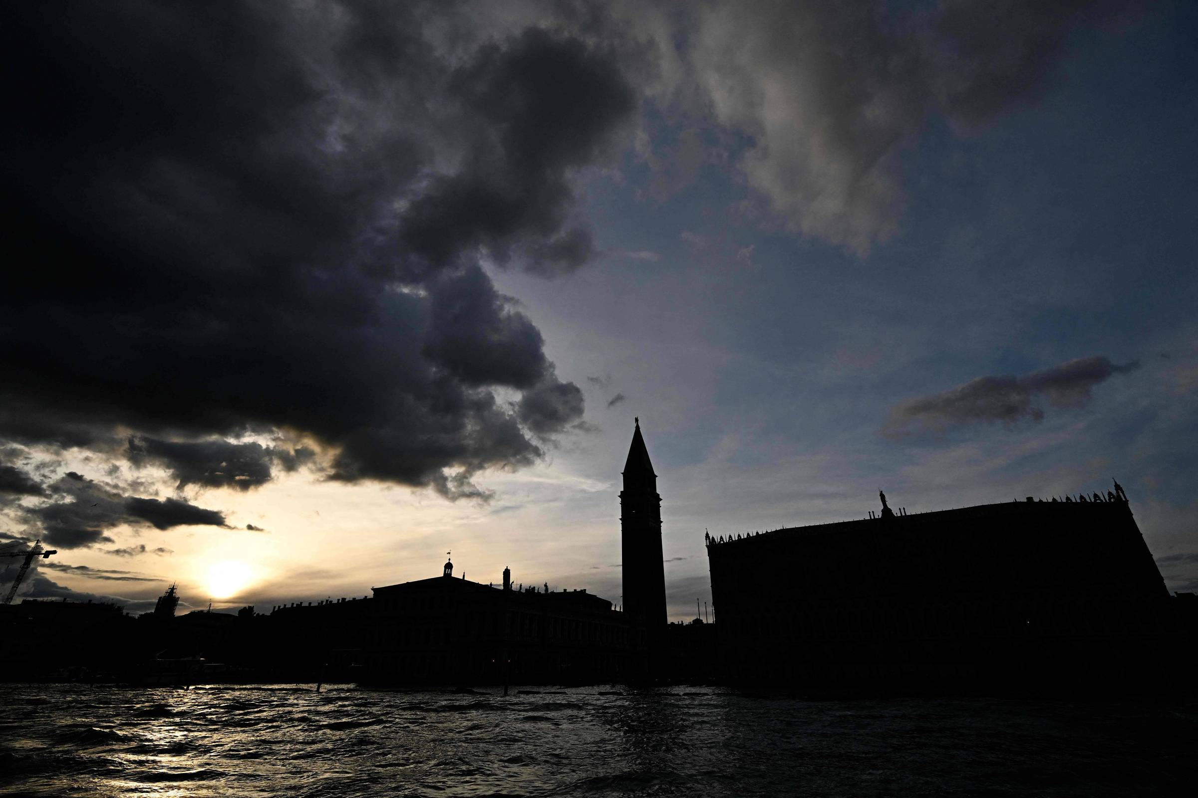 Silhueta do Campanário de São Marcos e edifícios adjacentes em Veneza contra um céu dramático com nuvens escuras ao pôr do sol, refletindo na água do canal.