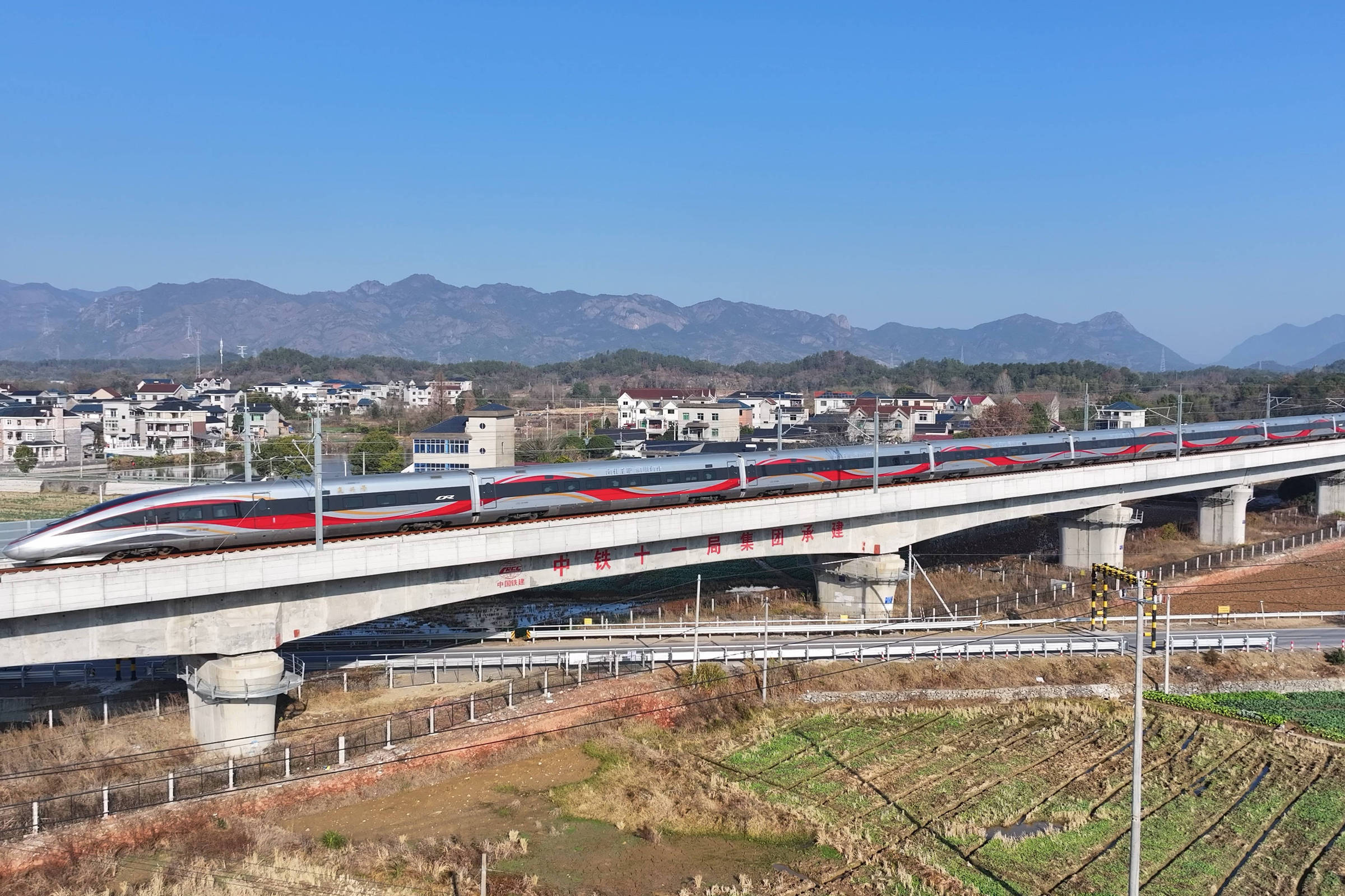Trem-bala branco com faixa vermelha circula sobre viaduto elevado em área rural. Casas e montanhas aparecem ao fundo sob céu azul claro.