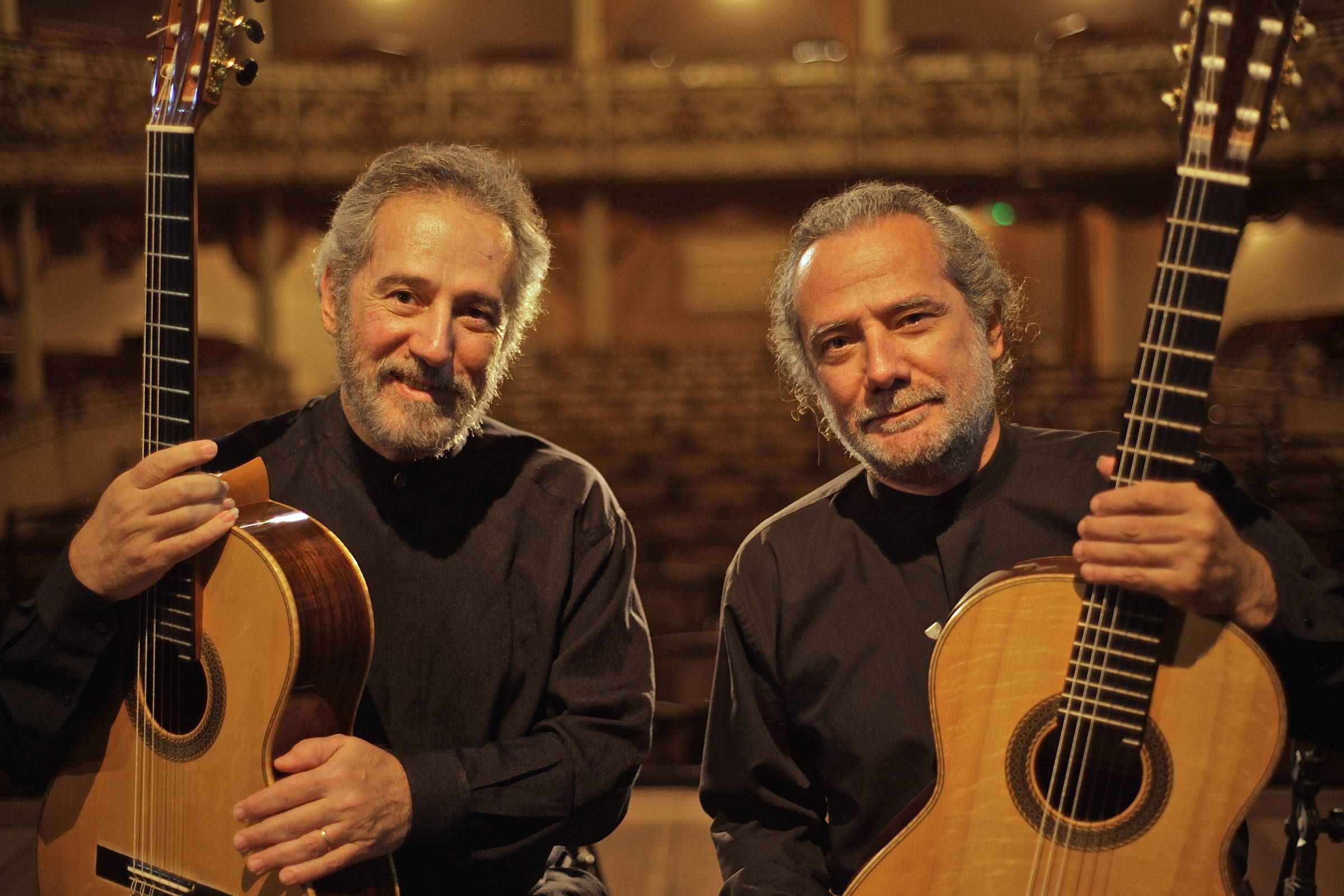 Dois homens estão posando para a foto em um ambiente de teatro. Ambos seguram violões de madeira clara. Eles estão vestidos com camisetas pretas e têm expressões amigáveis. Ao fundo, é possível ver assentos de um auditório.
