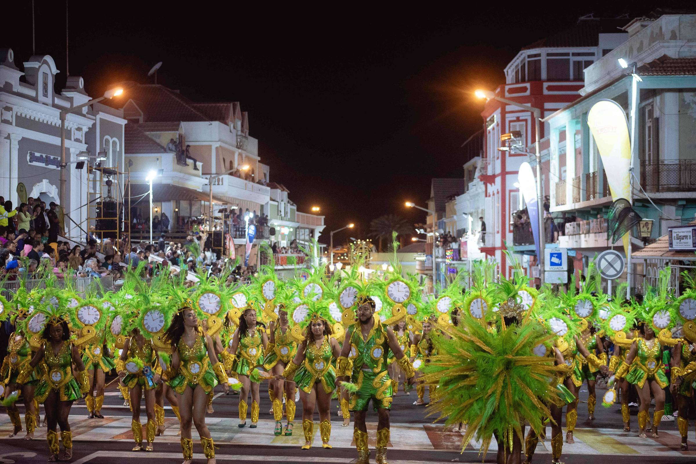 Grupo grande de pessoas desfilando à noite em rua urbana, vestindo fantasias verdes e douradas com adereços de penas e relógios decorativos. Edifícios iluminados e público assistindo nas calçadas ao fundo.