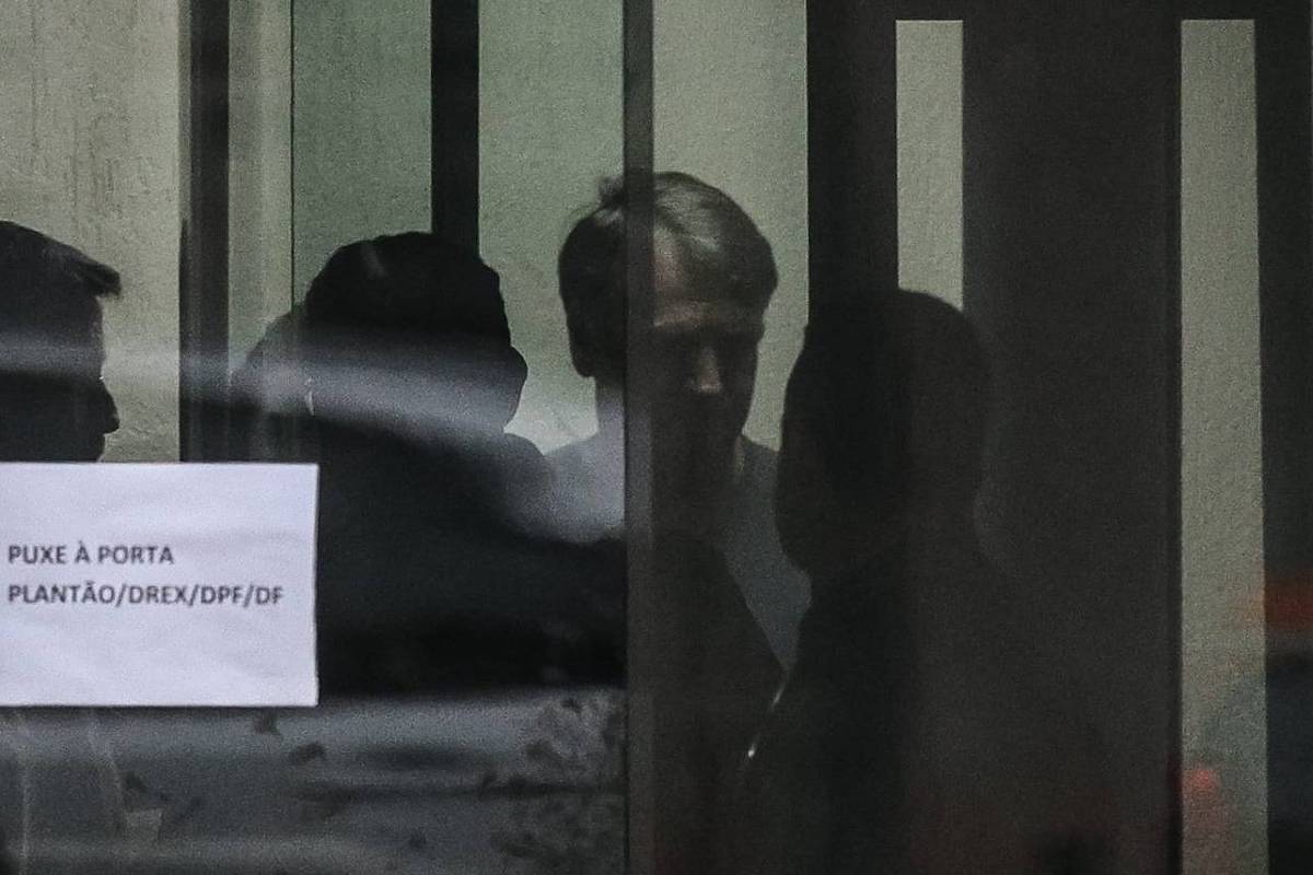 Quatro pessoas aparecem em silhueta atrás de uma porta de vidro com barras verticais. Na porta, um aviso branco com texto em português instrui a puxar a porta. A iluminação é baixa, dificultando a identificação dos rostos.