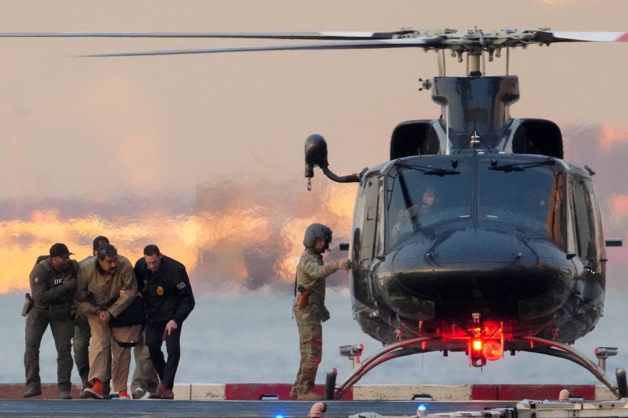 Quatro militares escoltam um homem algemado em direção a um helicóptero preto estacionado em plataforma. Um soldado com capacete está próximo à porta do helicóptero. O céu ao fundo apresenta tons alaranjados do pôr do sol.