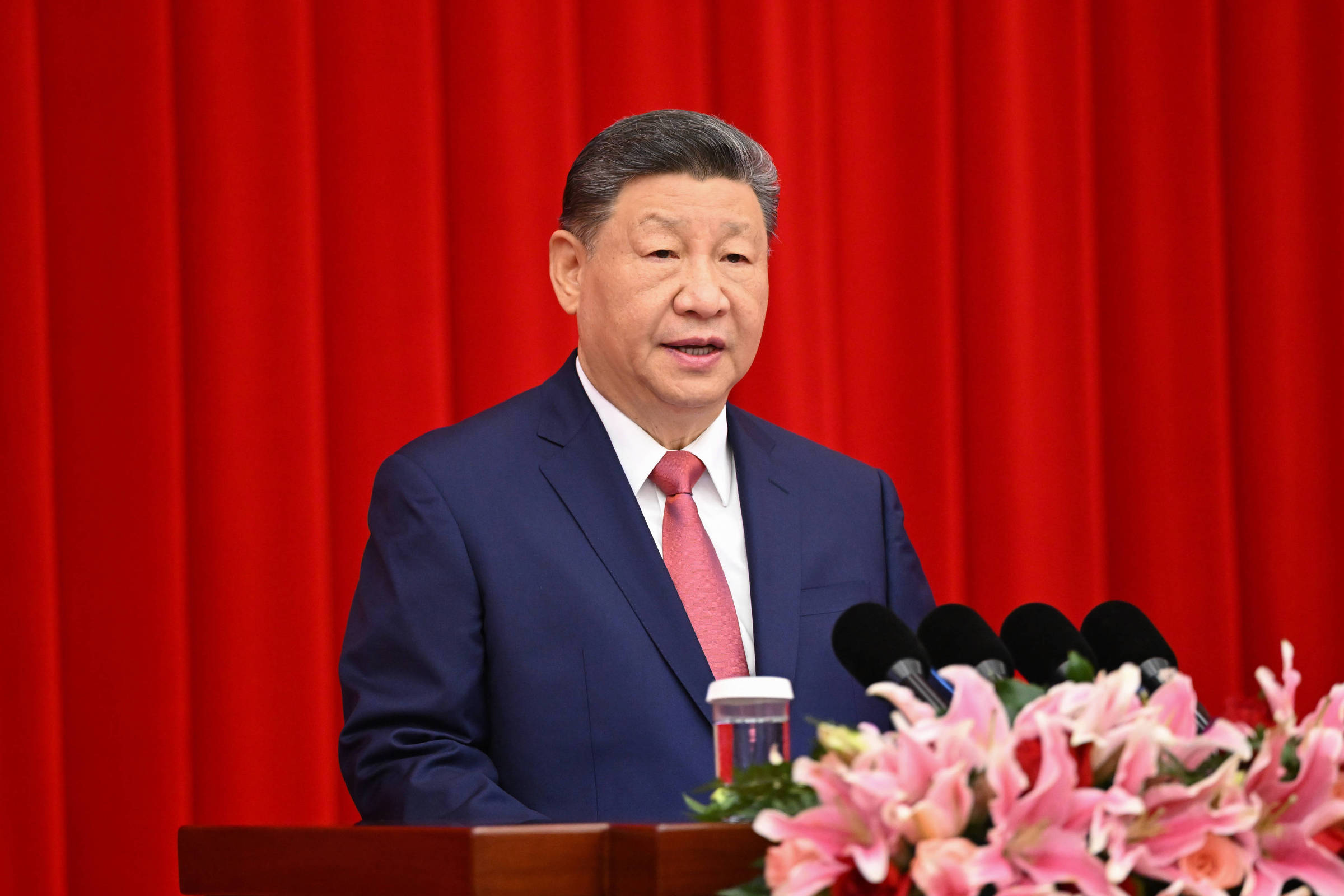 Homem de terno azul e gravata vermelha fala em púlpito com microfones, diante de cortina vermelha. Arranjo de flores rosas está à frente do púlpito.