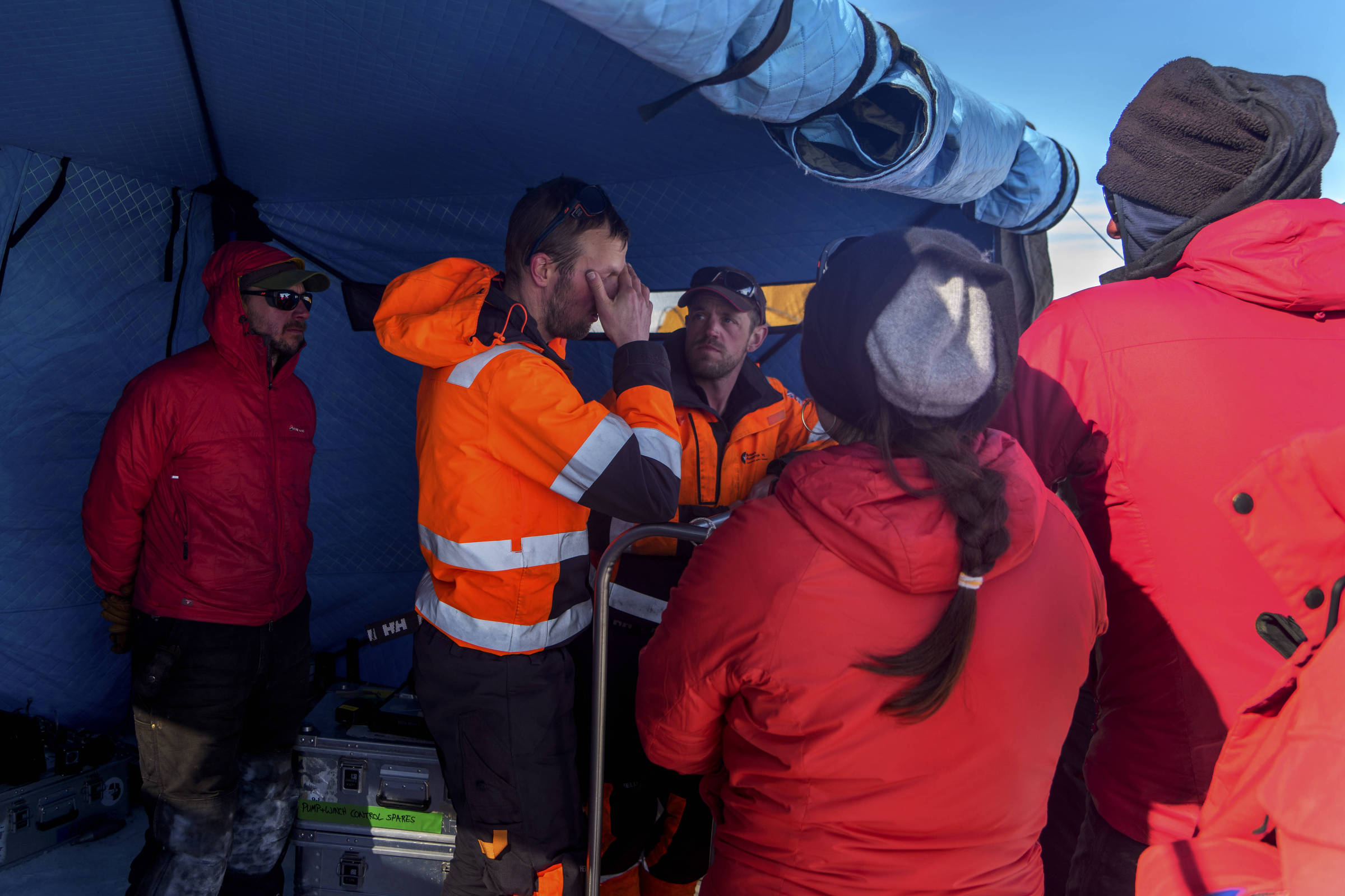 Seis pessoas vestindo roupas de frio, incluindo jaquetas vermelhas e coletes laranja com faixas refletivas, estão reunidas dentro de uma tenda azul. Um homem no centro, com colete laranja, coloca a mão sobre os olhos enquanto os demais o observam. Equipamentos e caixas estão no chão da tenda.