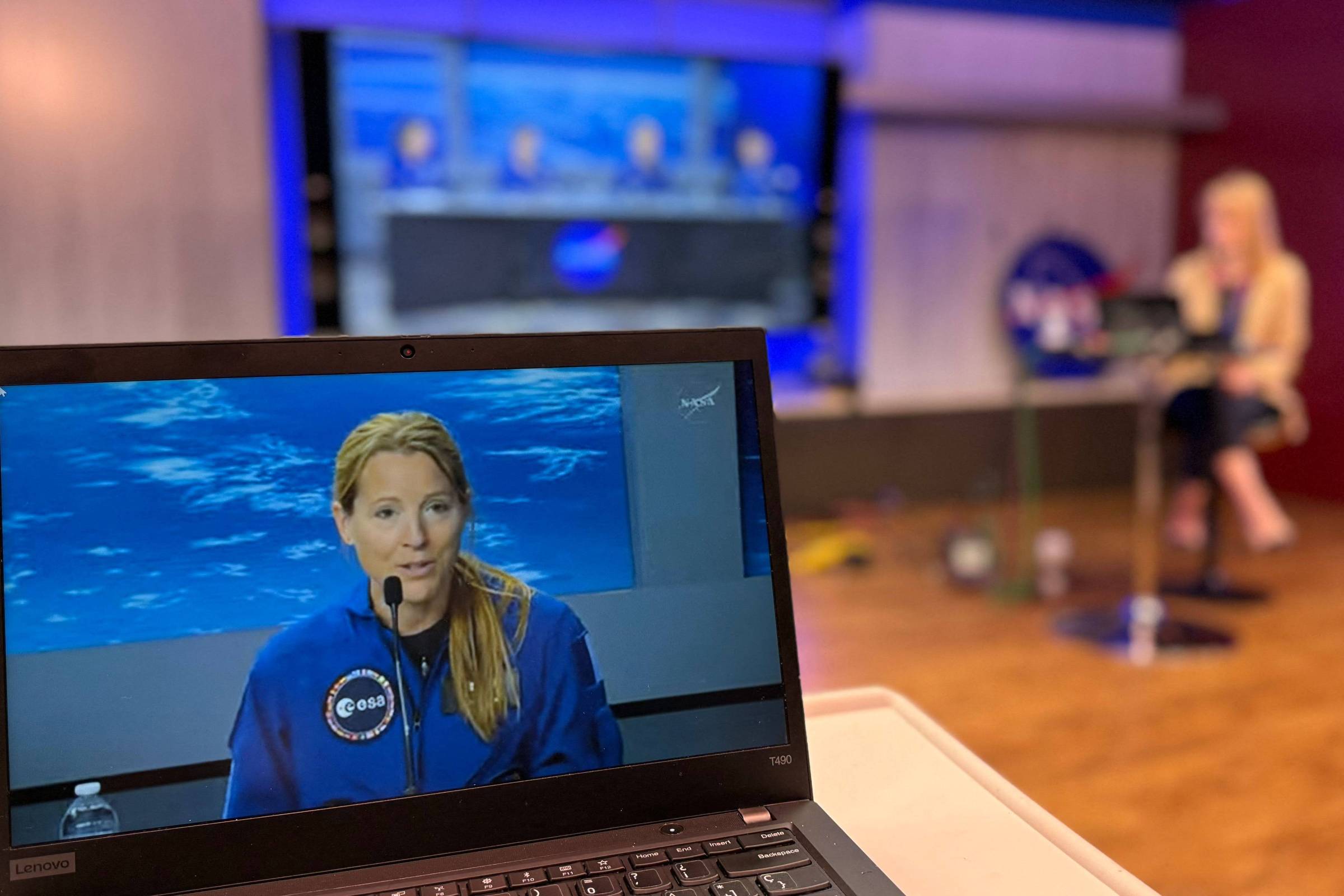 Astronauta aparece em vídeo na tela de um laptop, vestindo uniforme azul com emblema da ESA, falando em microfone. Ao fundo, mulher loira sentada em estúdio de TV com decoração azul e vermelha.