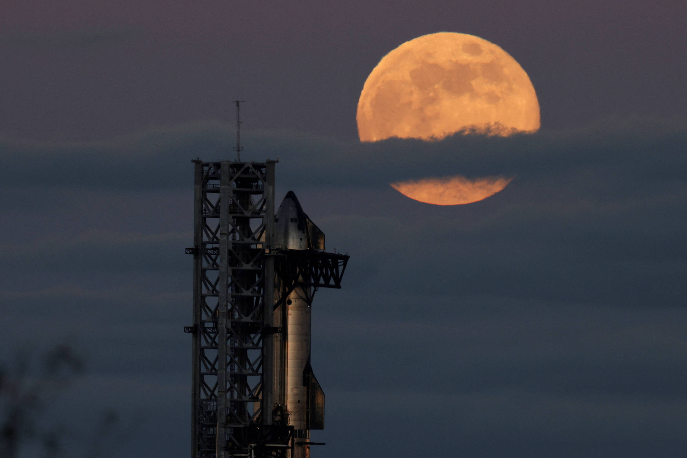 Foguete posicionado em plataforma de lançamento à esquerda, com grande lua cheia parcialmente encoberta por nuvens ao fundo, céu escuro ao entardecer.