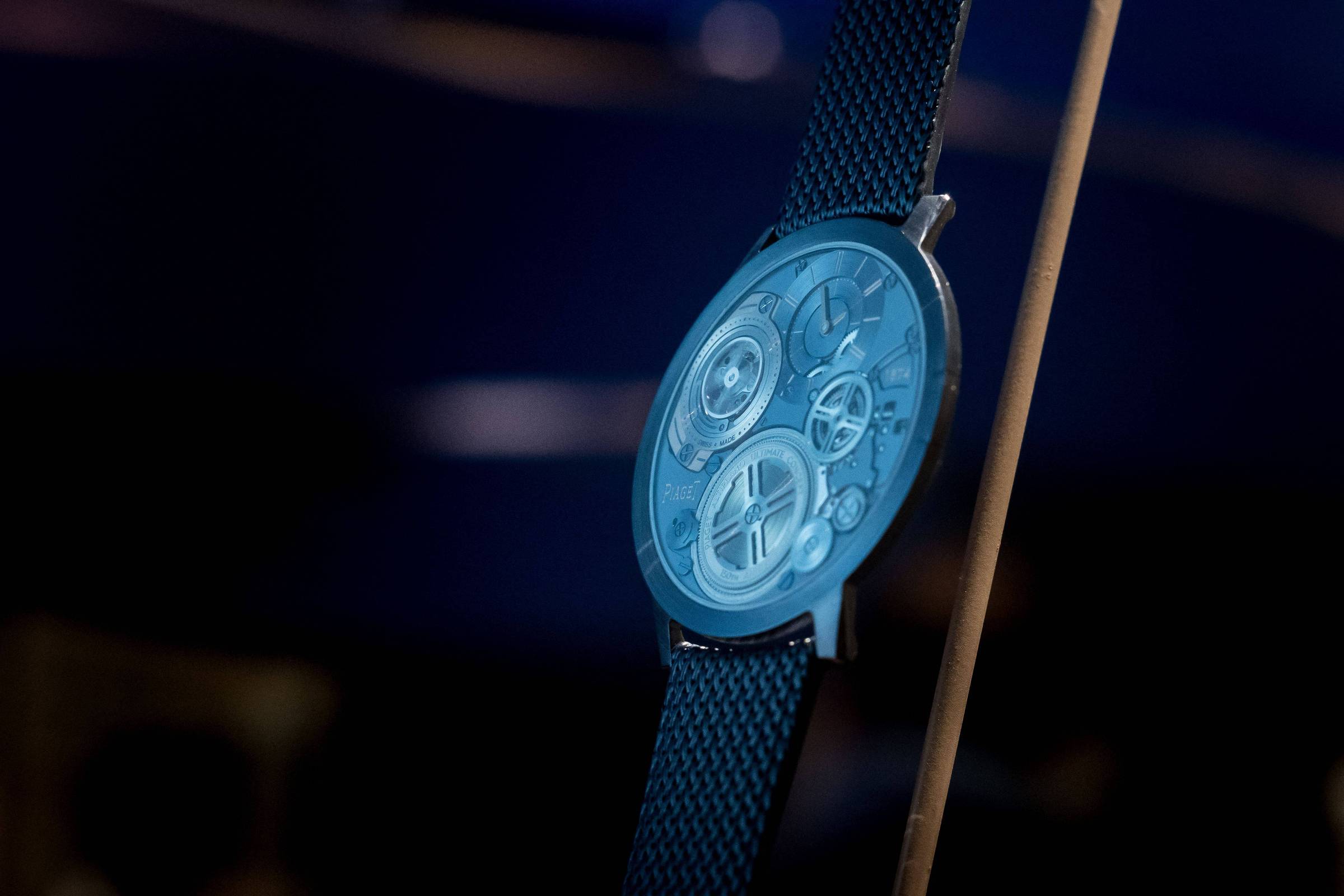 Relógio de pulso com mostrador transparente que revela engrenagens internas em tom azul. Pulseira preta de malha metálica, fundo escuro desfocado.