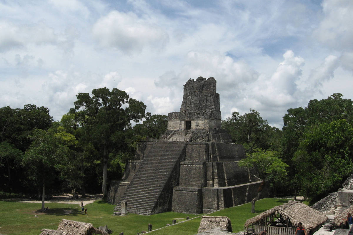 Pirâmide maia de pedra com escadaria central em sítio arqueológico de Tikal, Guatemala, cercada por árvores e céu parcialmente nublado.