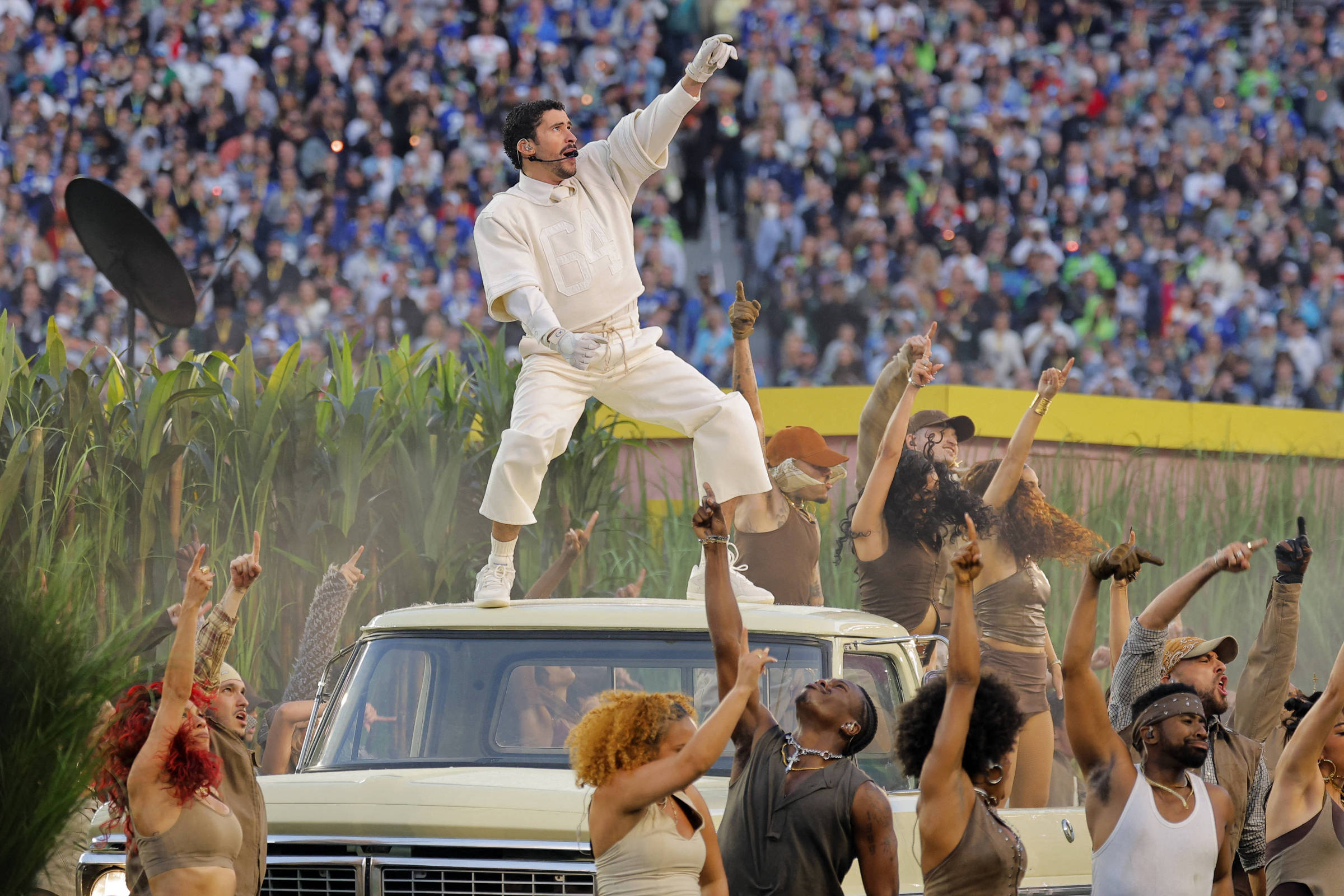 Artista vestido de branco está em pé sobre o capô de um veículo antigo, apontando para cima com a mão direita. Ao redor, grupo diversificado de pessoas com roupas em tons terrosos dança e levanta os braços. Ao fundo, multidão grande em arquibancada e vegetação alta