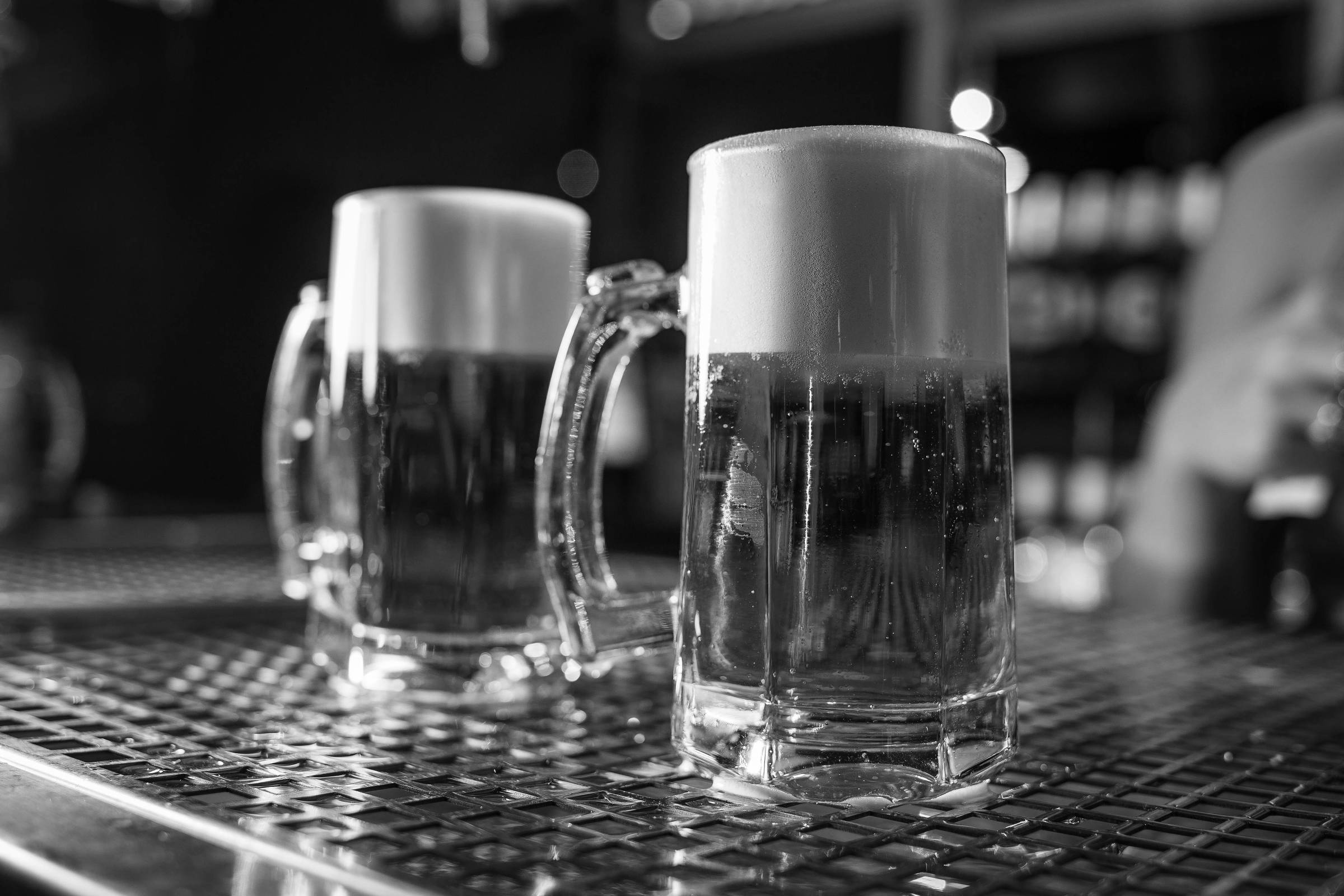 Duas canecas de cerveja com espuma no topo, colocadas sobre uma superfície metálica. A cerveja é de cor dourada e translúcida, e ao fundo, há uma leve desfocagem que sugere um ambiente de bar.